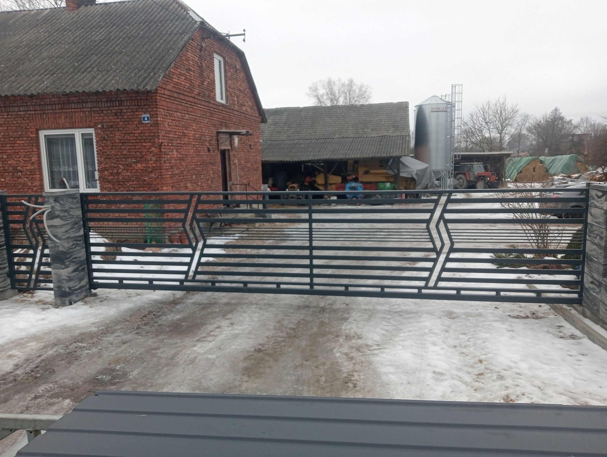 Nowoczesna, pozioma brama metalowa w kolorze grafitowym z geometrycznym wzorem na tle zimowego krajobrazu wiejskiego z budynkami gospodarczymi i traktorem.