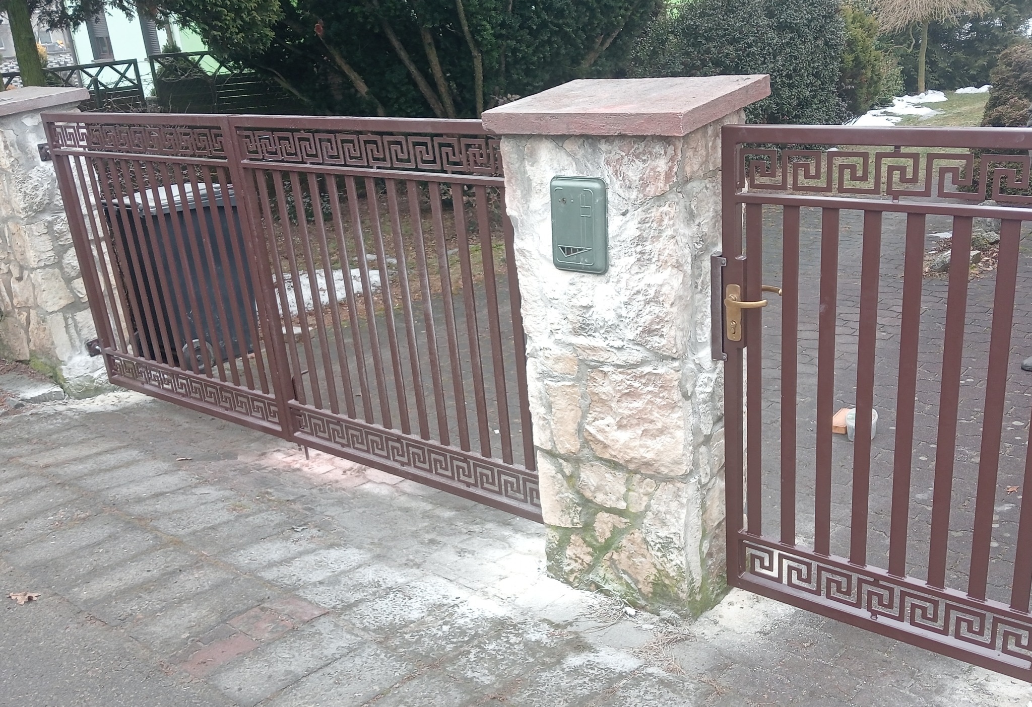 Brama wjazdowa z metalu, pomalowana na brązowo, z dekoracyjnym wzorem greckim, osadzona na kamiennych słupkach z zieloną skrzynką na listy.