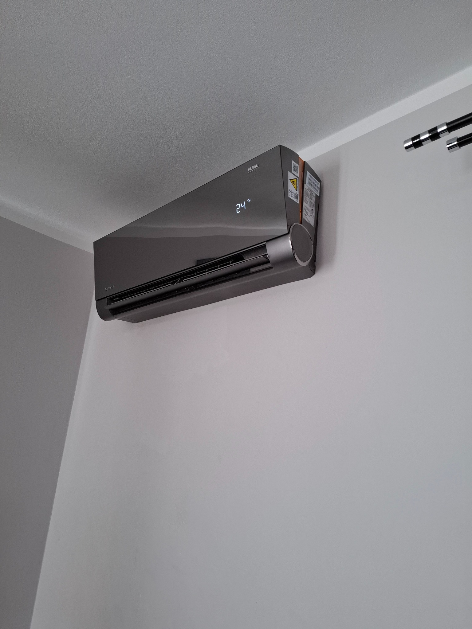 Czarny klimatyzator ścienny Versu z wyświetlaczem temperatury 24 stopnie Celsjusza zamontowany w rogu pomieszczenia z białymi ścianami.