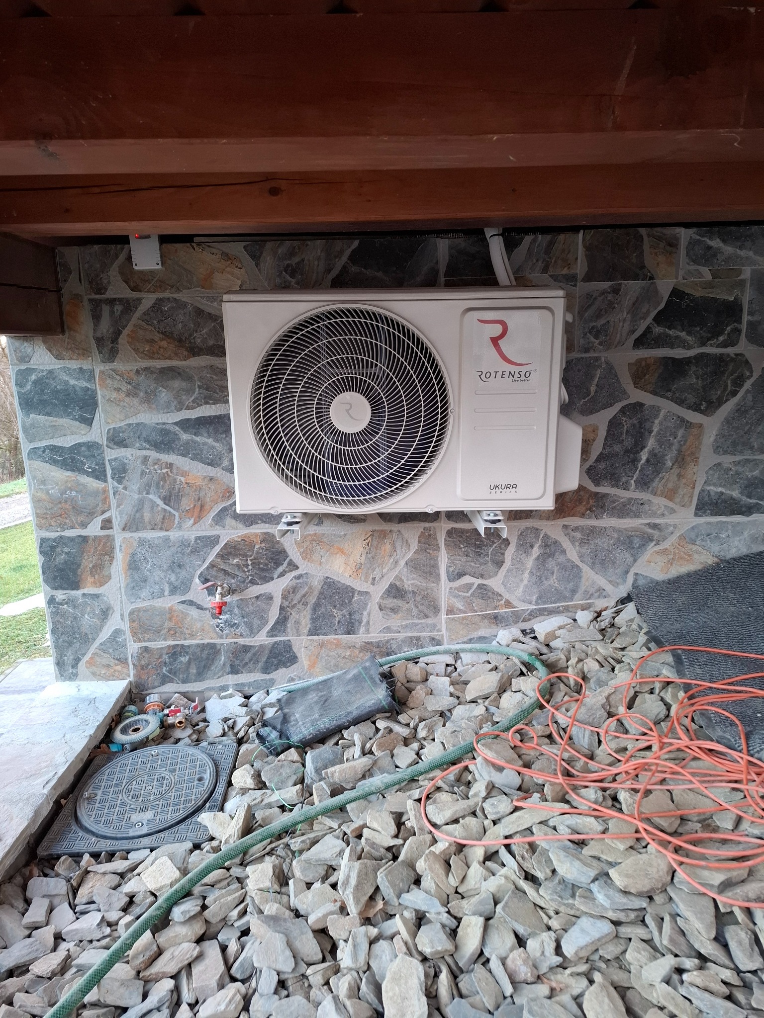Zewnętrzna jednostka klimatyzatora marki Rotenso zamontowana na ścianie z kamienia dekoracyjnego, widoczne przewody i drobne kamienie na podłożu.