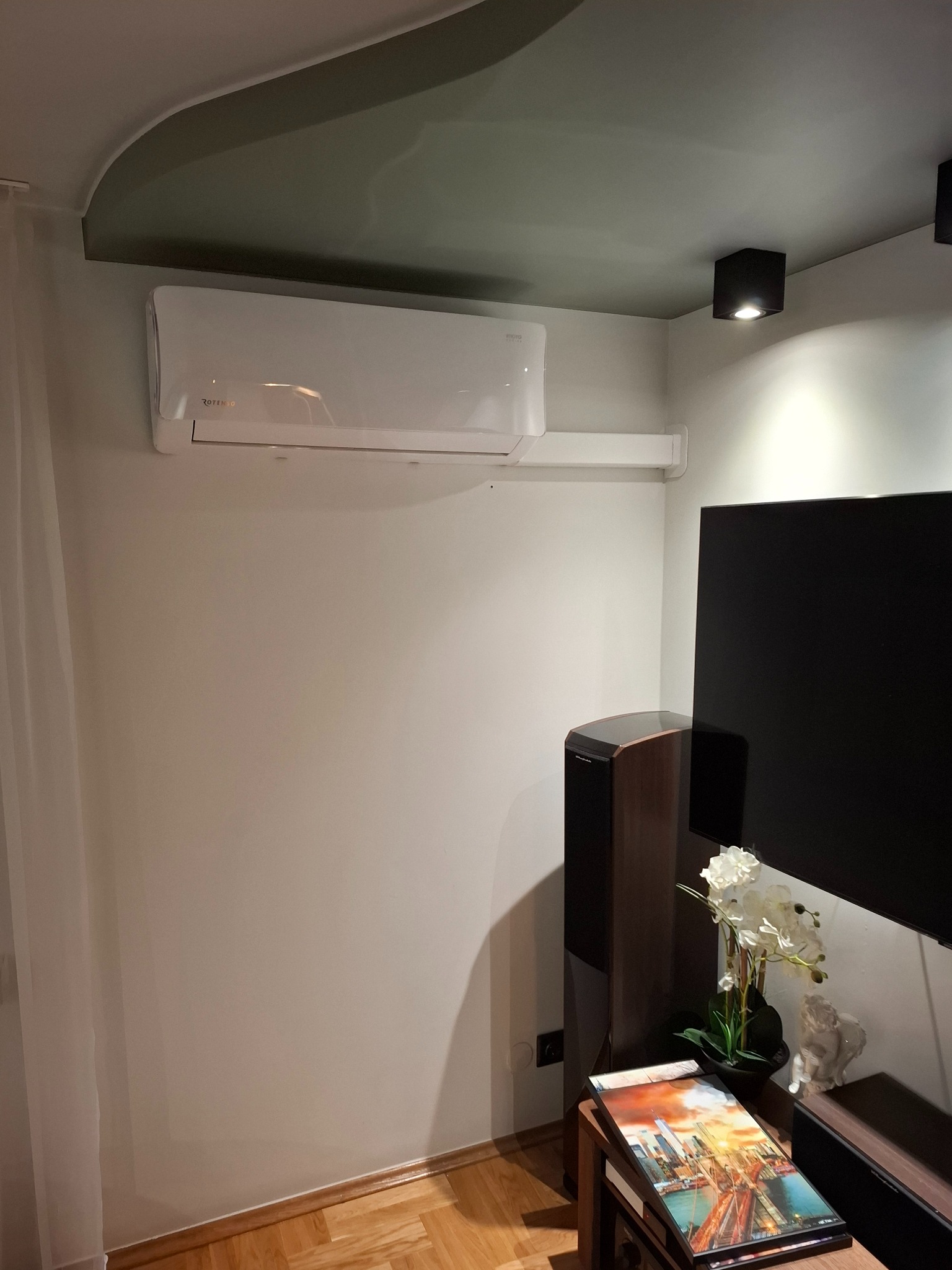 Biała jednostka klimatyzacji Rotenso zamontowana na ścianie w nowoczesnym salonie, widoczny głośnik, telewizor i dekoracje na szafce RTV.