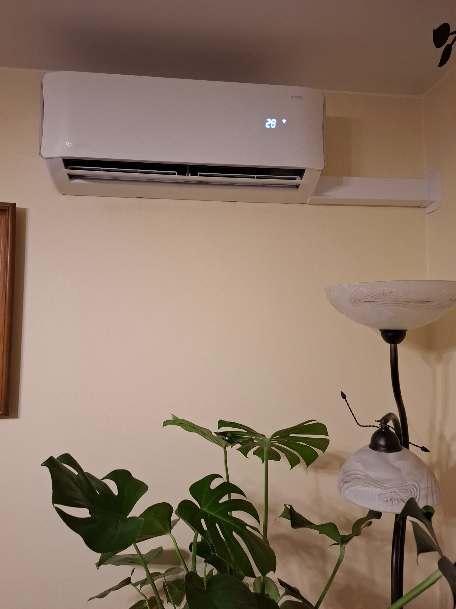 Biała klimatyzacja ścienna marki Rotenso Imoto Series z wyświetlaną temperaturą 28 stopni, zamontowana w jasnym pomieszczeniu z rośliną Monstera i lampą podłogową.