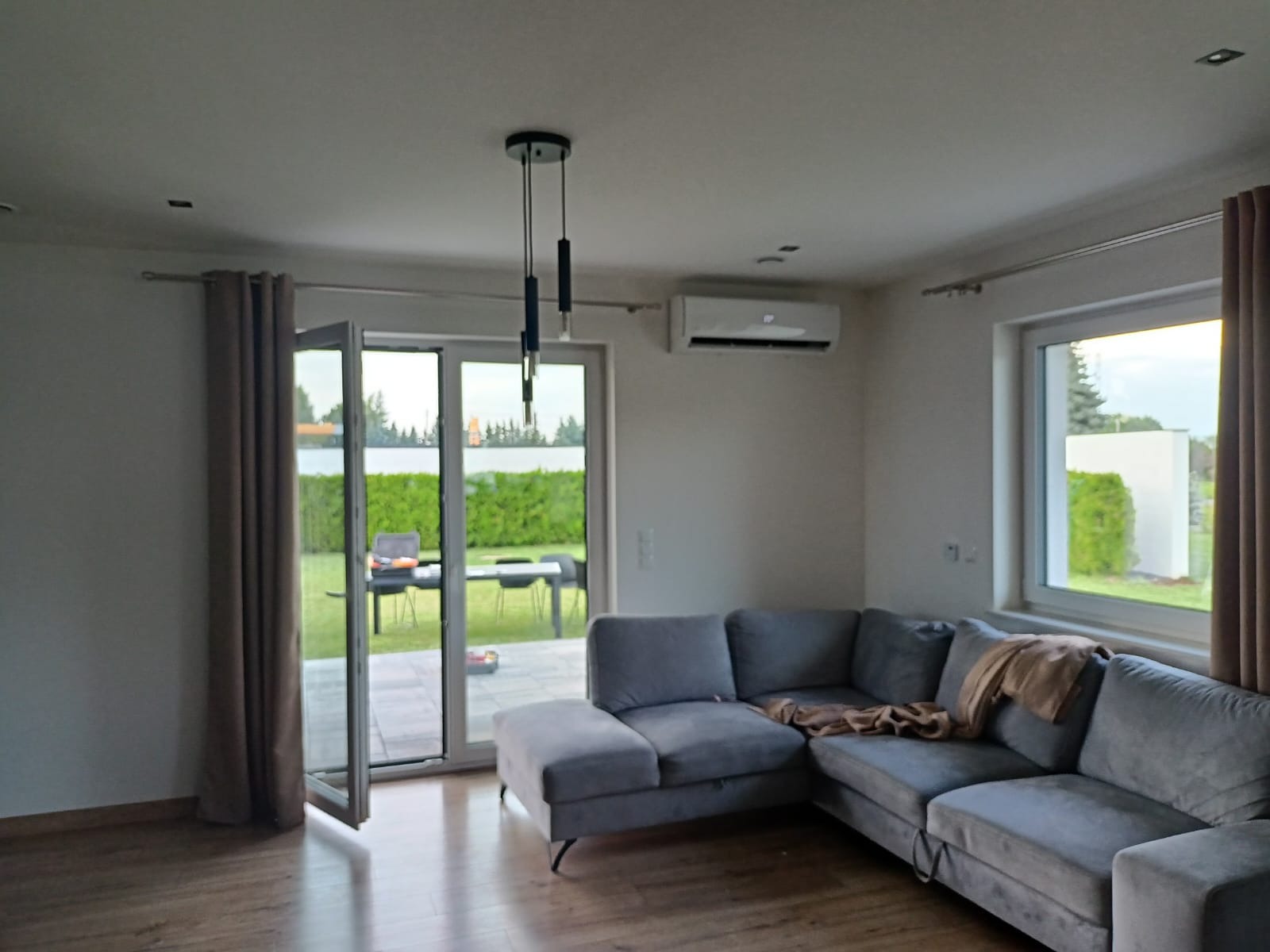 Nowoczesne wnętrze salonu z szarą sofą narożną i klimatyzatorem ściennym zamontowanym nad drzwiami balkonowymi, widok na ogród