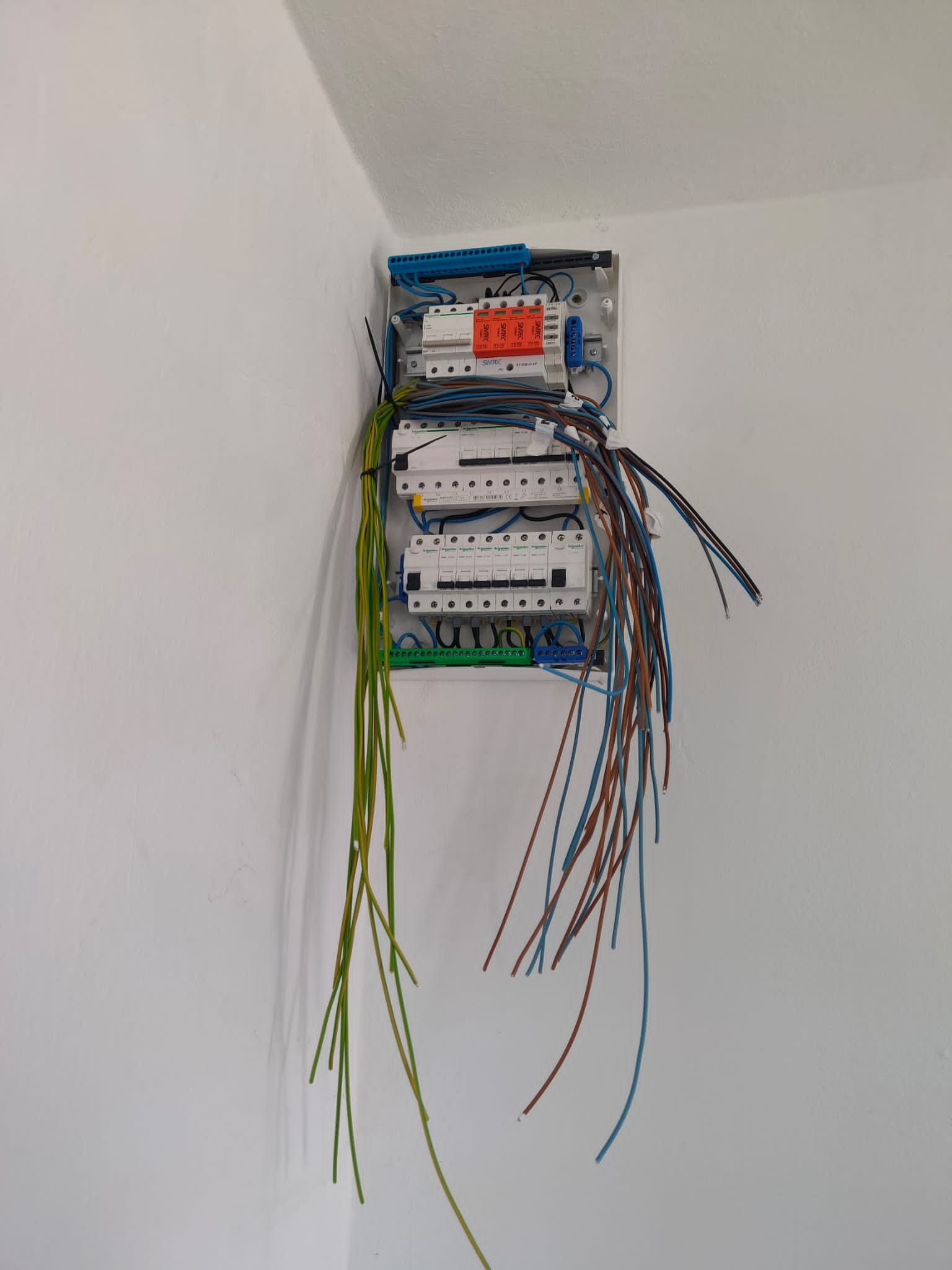 Rozdzielnica elektryczna z widocznymi przewodami w trakcie montażu na ścianie, z dużą ilością wystających kabli w kolorach brązowym, niebieskim i zielono-żółtym.