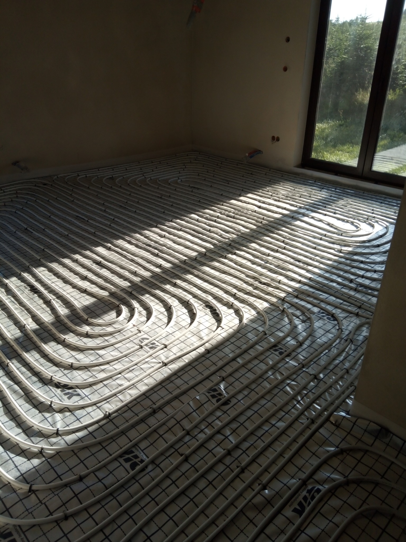 Białe rury ogrzewania podłogowego ułożone spiralnie na folii z nadrukowaną kratką w jasnym pokoju z dużym oknem, fragmenty podłogi oświetlone promieniami słonecznymi.