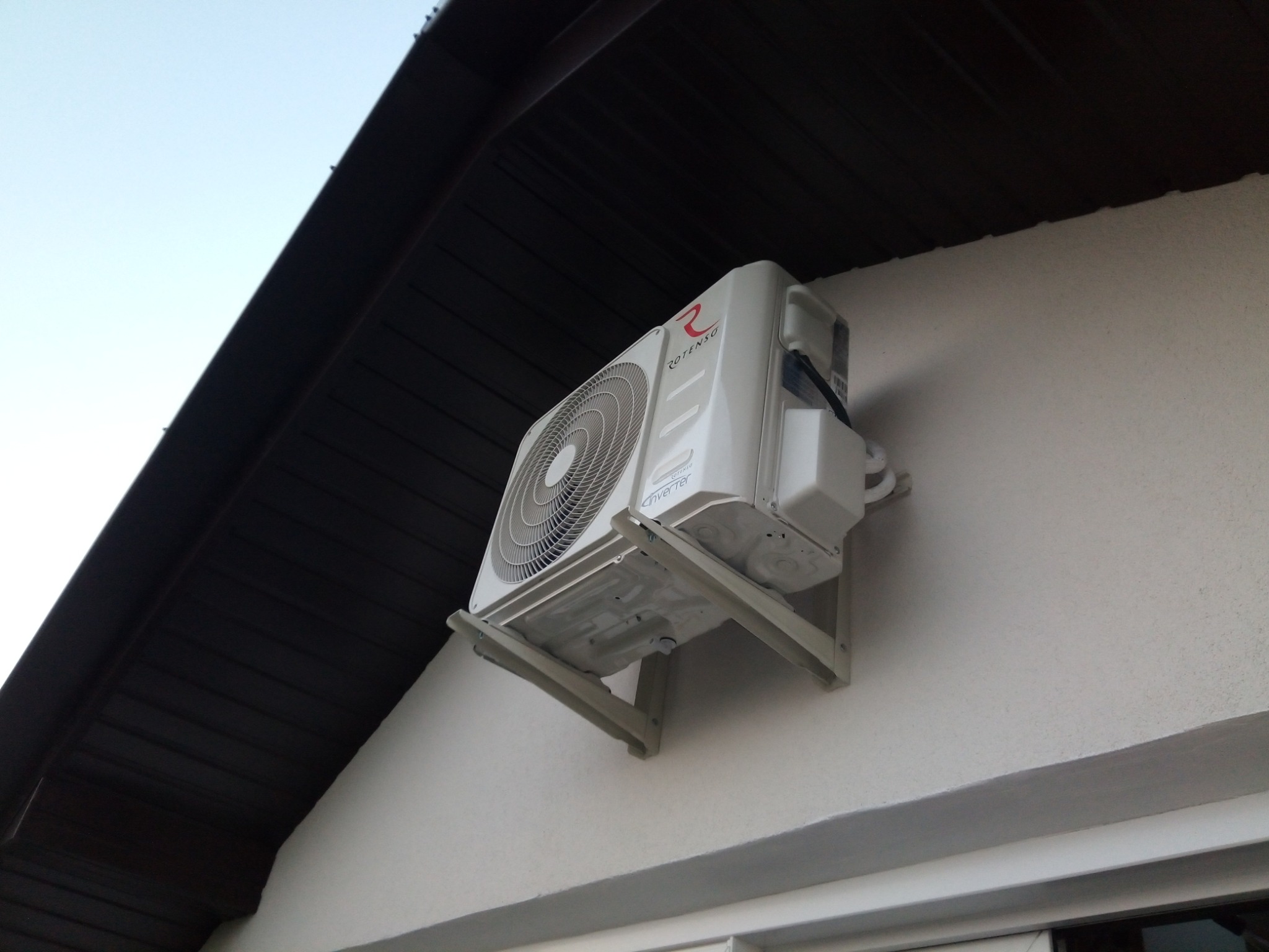 Zewnętrzna jednostka klimatyzatora Rotenso zamontowana na ścianie domu pod okapem dachu, widoczna z dołu pod kątem.