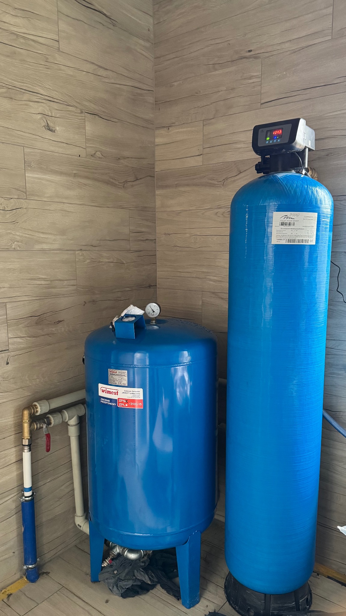 Niebieski zestaw hydroforowy z manometrem i elektronicznym sterownikiem, ustawiony w rogu pomieszczenia wykończonego panelami imitującymi drewno.