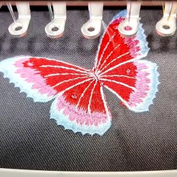 FASHIONPRO Gabriela Dobroń - Czerwono-biały motyl z różowymi i błękitnymi akcentami haftowany na ciemnoszarym materiale, widoczne elementy maszyny do haftowania.