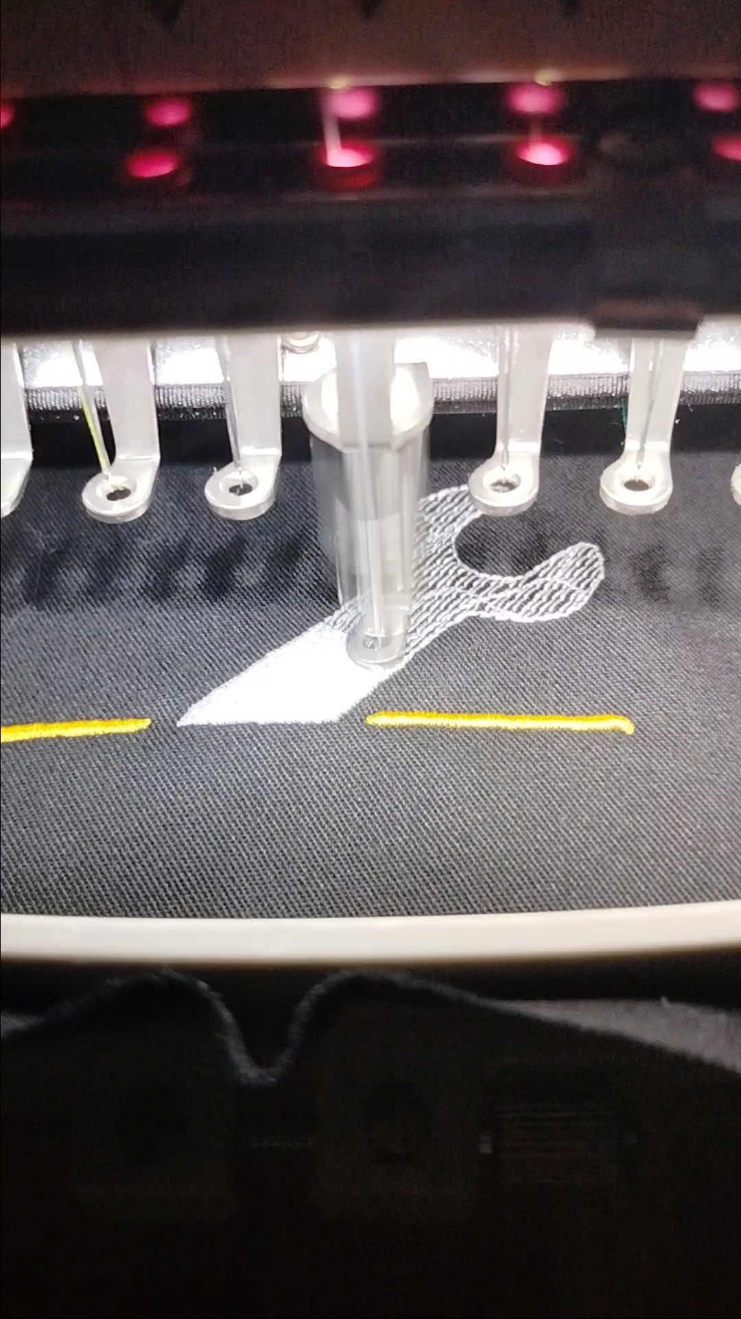 Proces tworzenia białego wzoru na ciemnoszarym materiale za pomocą maszyny, widoczne żółte detale i igła w trakcie pracy.