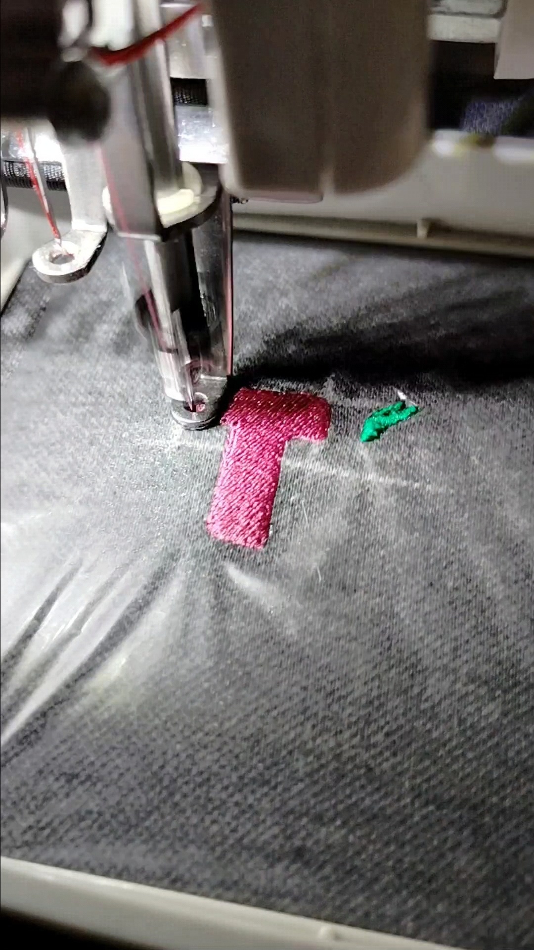 Maszyna do szycia wykonuje różowy haft w kształcie litery T na szarym materiale, obok widoczny fragment zielonego haftu.