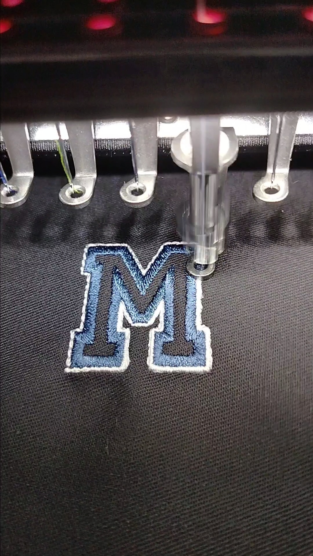 Proces tworzenia haftu komputerowego: igła maszyny precyzyjnie wyszywa literę 'M' na ciemnym materiale, tworząc niebiesko-biały wzór z wyraźną teksturą.