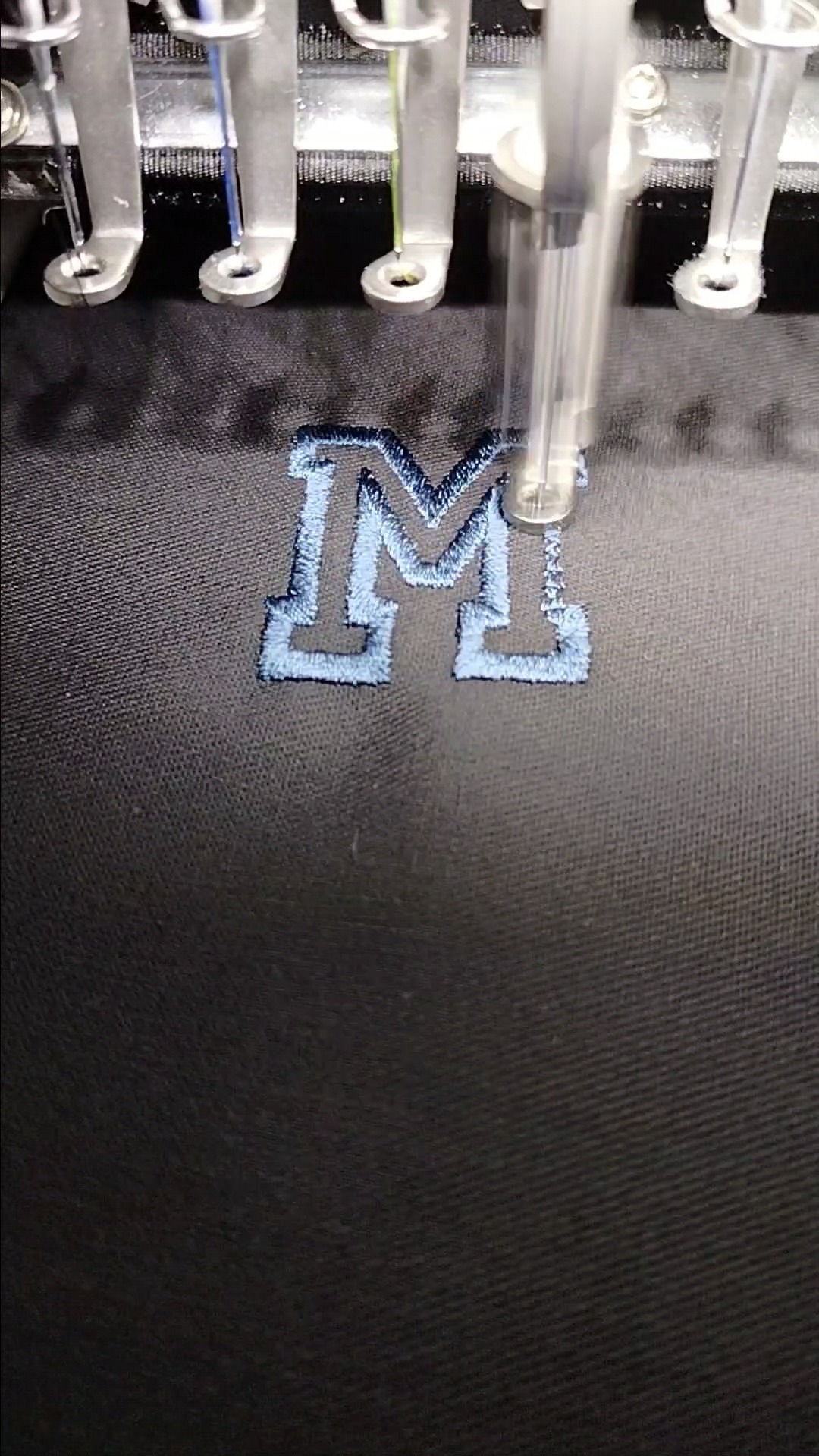 Proces tworzenia niebieskiego haftu litery M na ciemnej tkaninie za pomocą maszyny do haftowania z wieloma igłami.