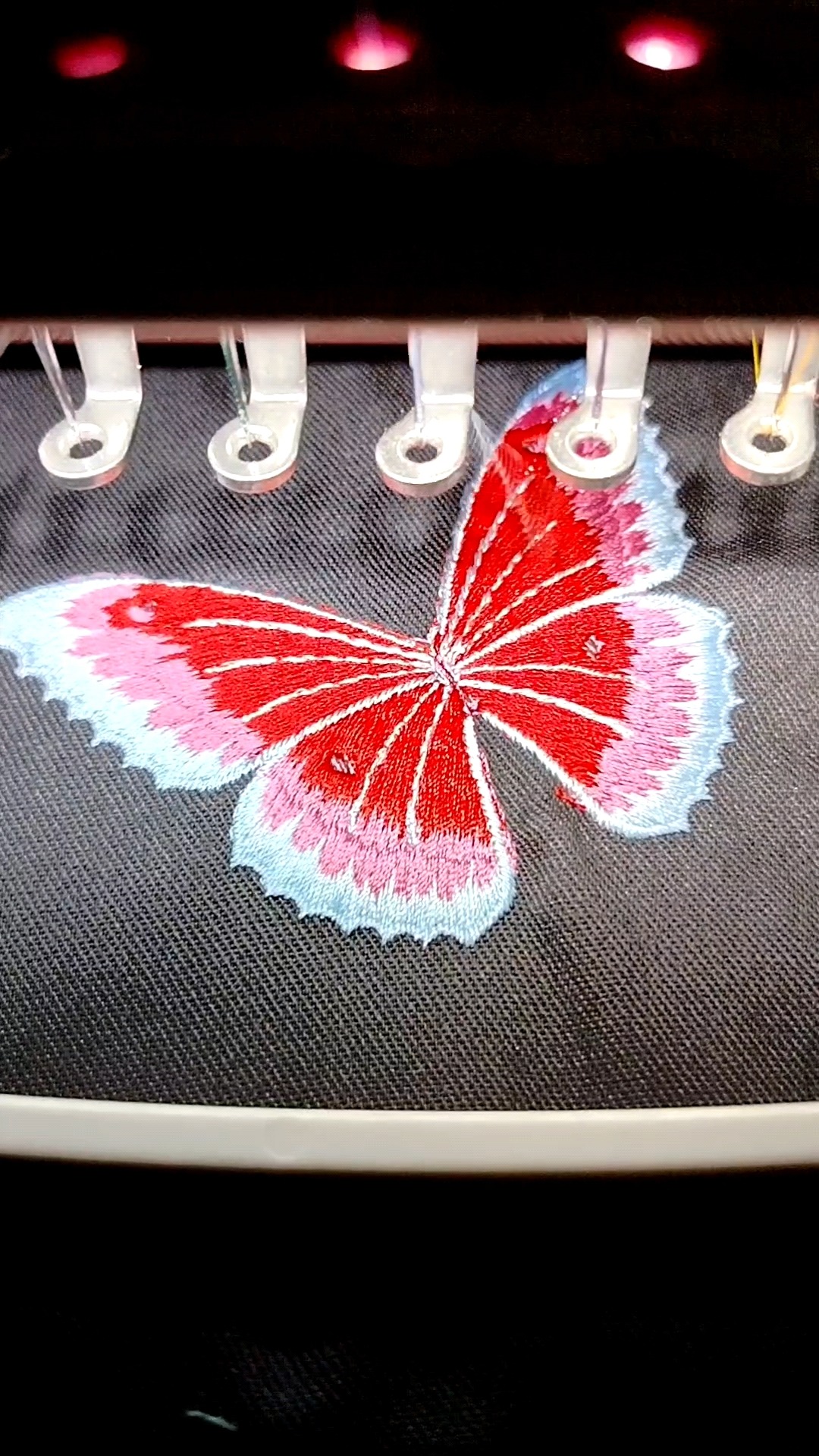 Czerwono-biały motyl z różowymi i błękitnymi akcentami haftowany na ciemnoszarym materiale, widoczne elementy maszyny do haftowania.
