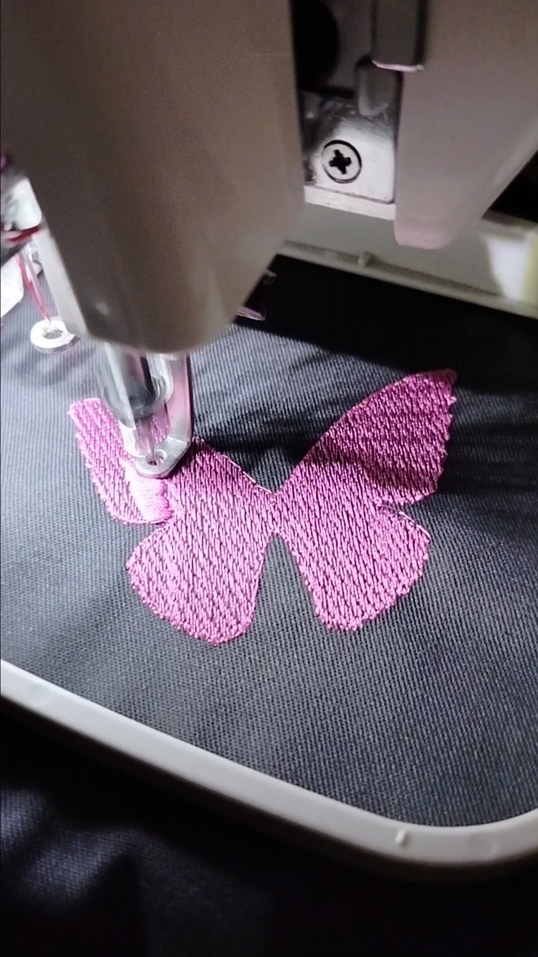 Proces haftowania różowego motyla na szarej tkaninie za pomocą maszyny do szycia.