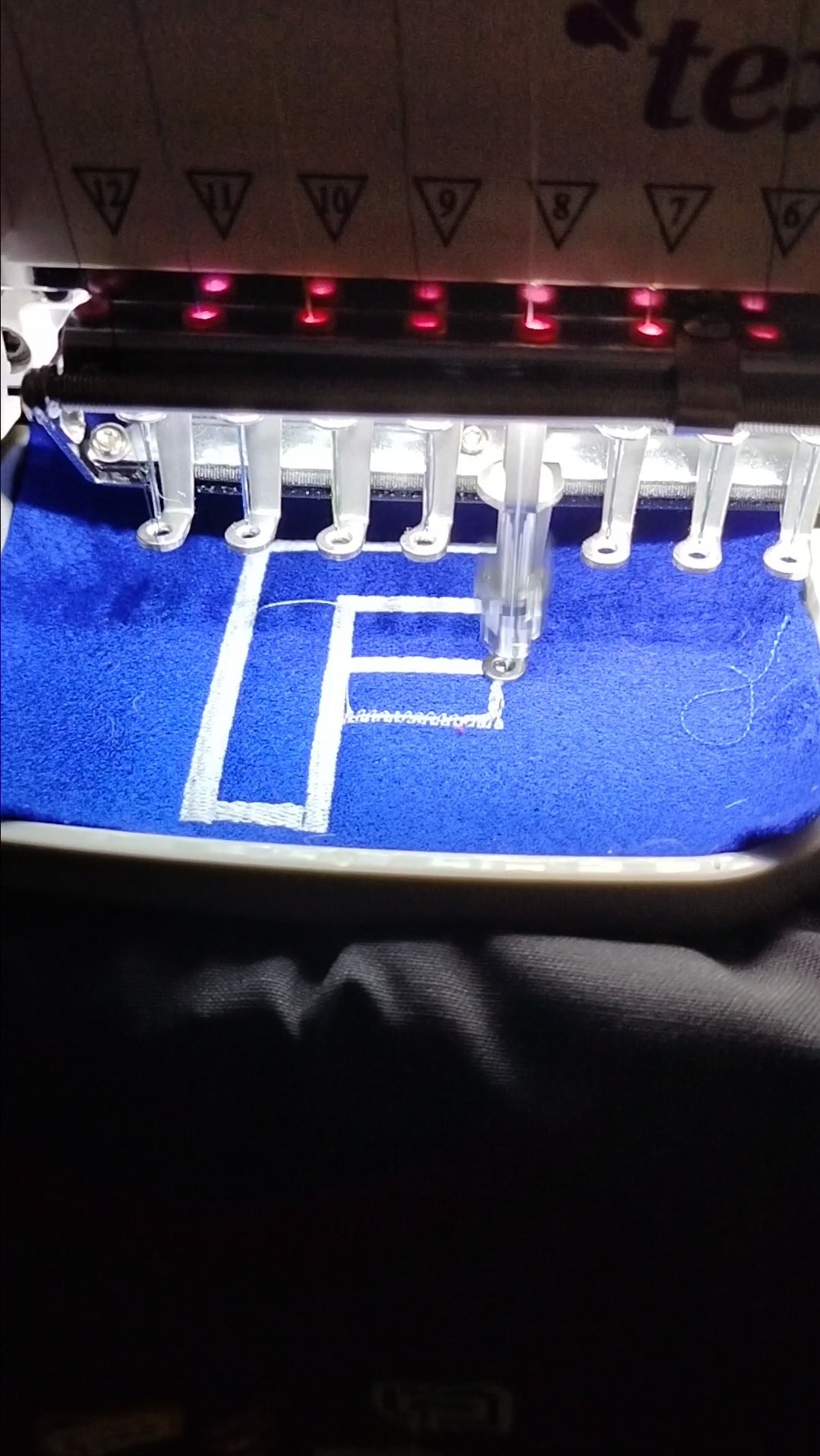 Proces tworzenia białego haftu litery 'F' na niebieskim materiale przy użyciu przemysłowej maszyny do haftowania z widocznymi igłami i elementami mocującymi materiał.