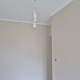 Bogusław Paszek - Jasny pokój po malowaniu z widoczną żarówką wiszącą na kablu i gładkimi ścianami w odcieniach beżu i bieli. Widoczny fragment listwy przysufitowej.