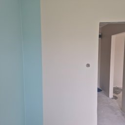 Bogusław Paszek - Ściana po malowaniu w odcieniach błękitu i beżu, widoczne gniazdko elektryczne i surowa podłoga w przejściu. Minimalistyczne wykończenie wnętrza.