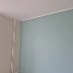 Pomieszczenie po malowaniu: ściany w odcieniach szarości i błękitu, biały sufit z listwą. Dwie rury pionowo przy ścianie. Widok z rogu pokoju.