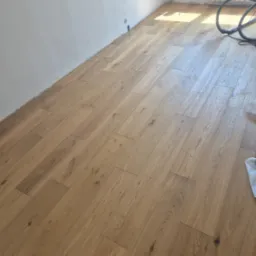 Jasne panele podłogowe ułożone w pokoju z białymi ścianami. Widoczny fragment grzejnika i rura od odkurzacza. Podłoga po ułożeniu, gotowa do użytku.