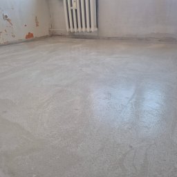 Bogusław Paszek - Wyrównana, szara wylewka podłogowa w pokoju z grzejnikiem i fragmentami starej farby na ścianach. Widoczne ślady po wałku na powierzchni.
