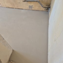 Bogusław Paszek - Wyrównana, świeżo wylana szara posadzka w rogu pomieszczenia z widocznymi rurami pod sufitem. Ściany w trakcie remontu.