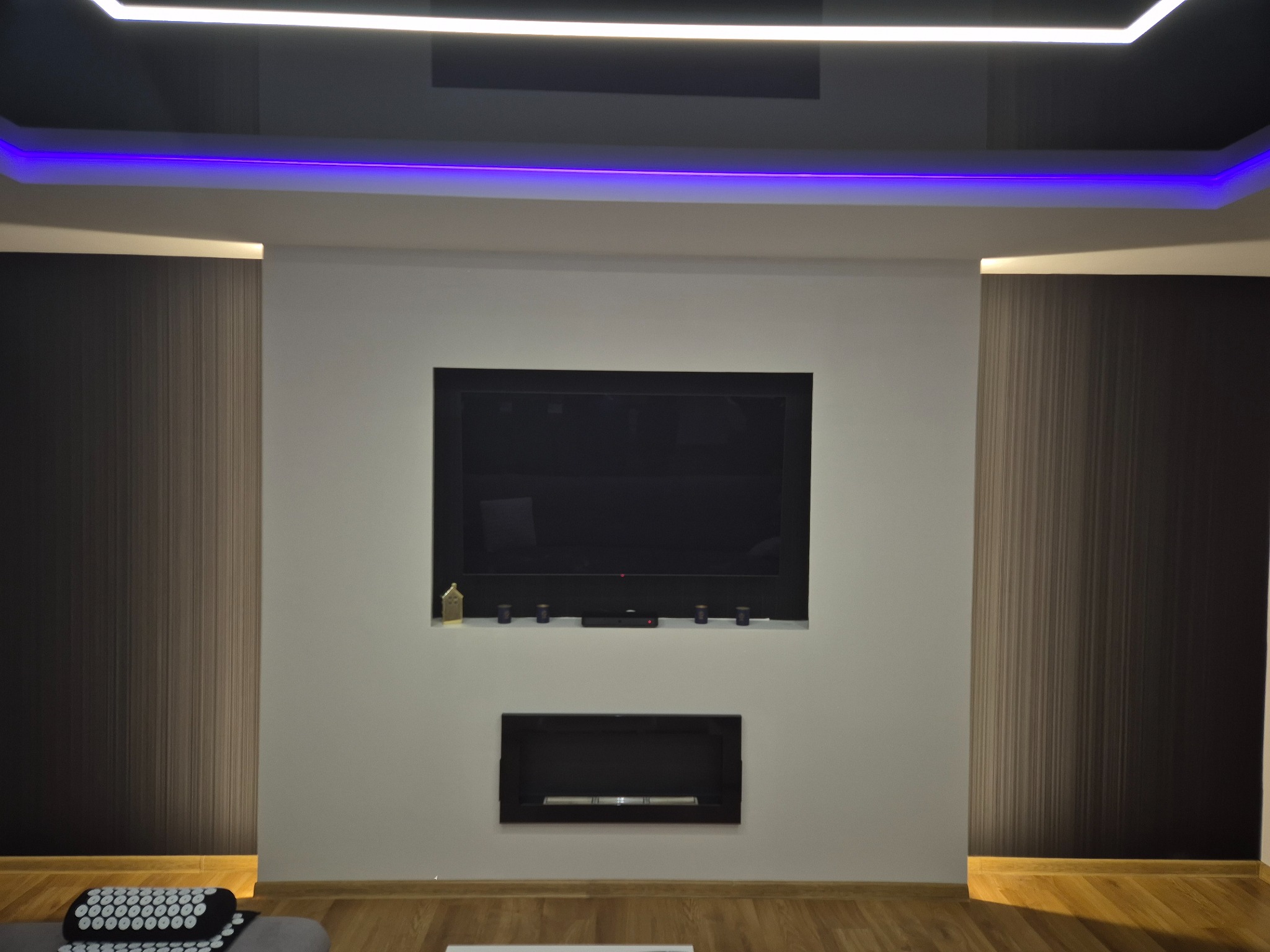 Nowoczesne wnętrze salonu z podwieszanym sufitem z oświetleniem LED, ściany wykończone tapetą w pionowe pasy i gładzią, wnęka z telewizorem i biokominkiem.