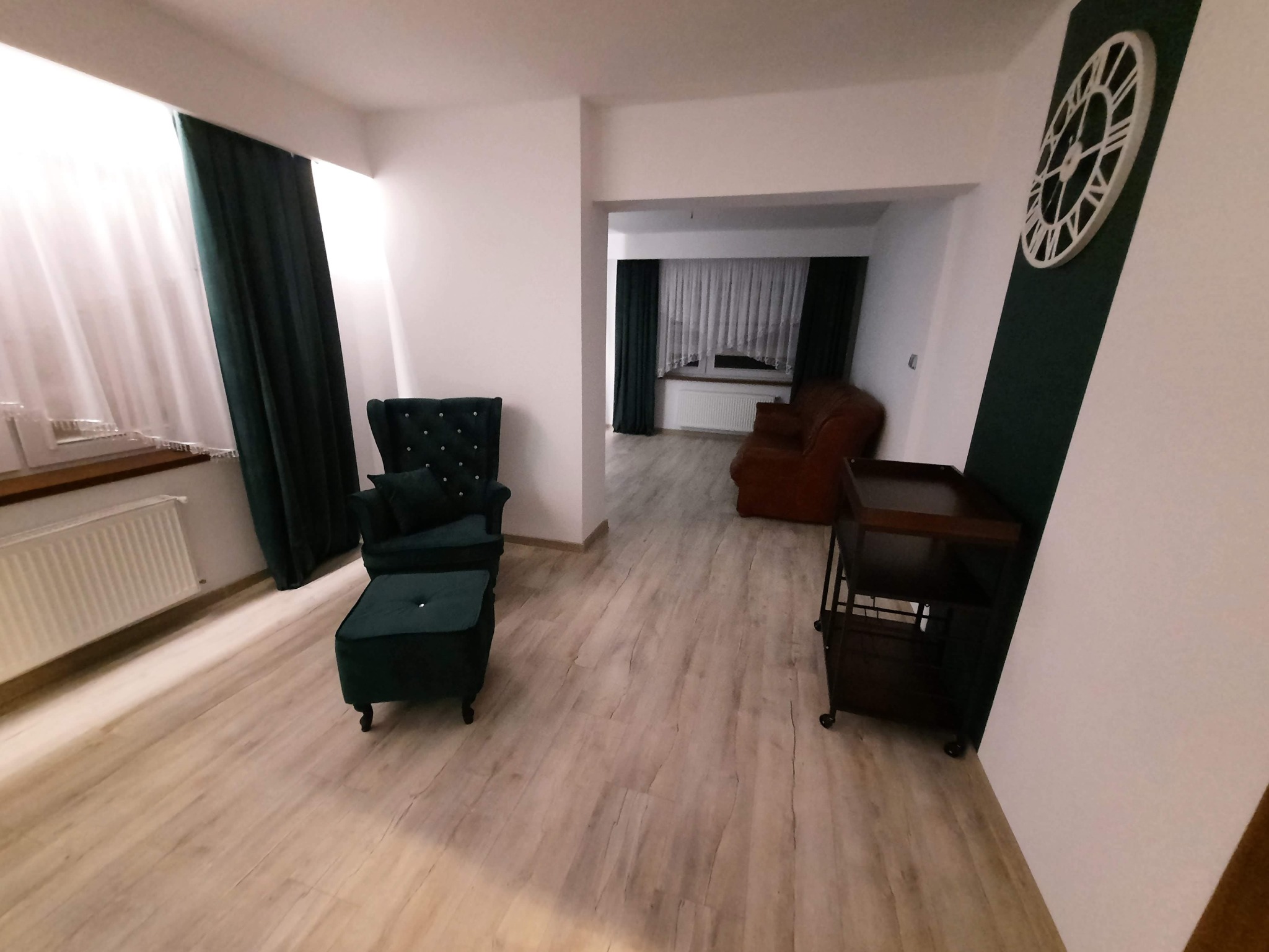 Salon z jasną podłogą, ciemnozielonymi zasłonami i tapicerowanym fotelem z podnóżkiem w tym samym kolorze. Na ścianie dekoracyjny zegar.