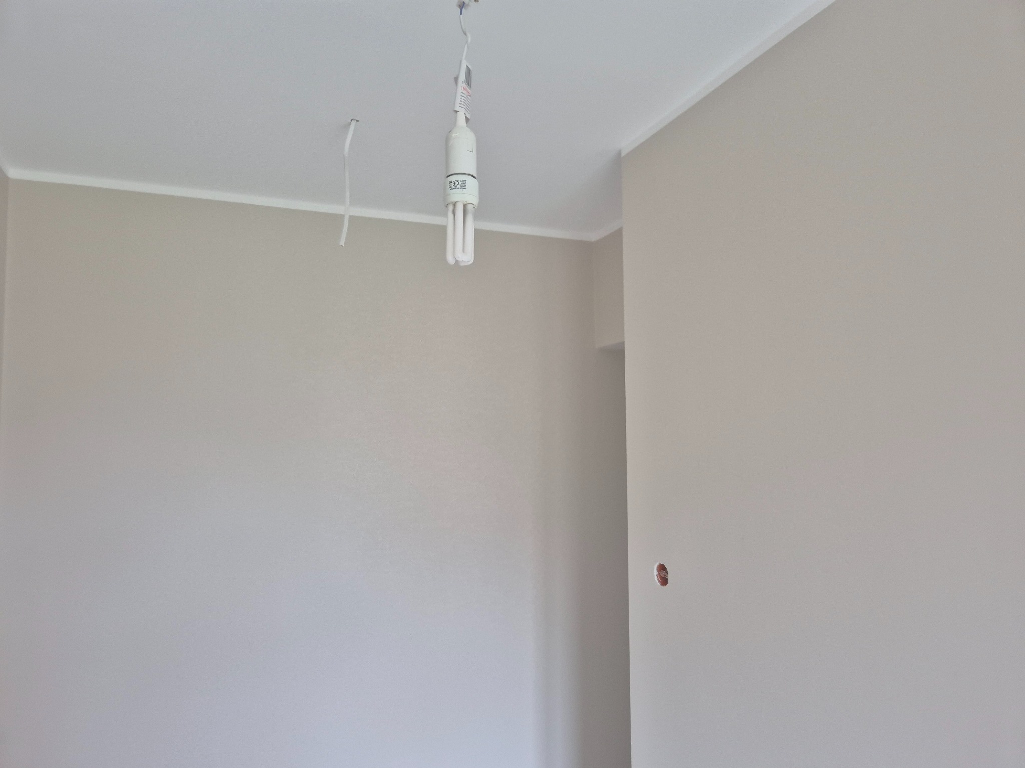 Jasny pokój po malowaniu z widoczną żarówką wiszącą na kablu i gładkimi ścianami w odcieniach beżu i bieli. Widoczny fragment listwy przysufitowej.