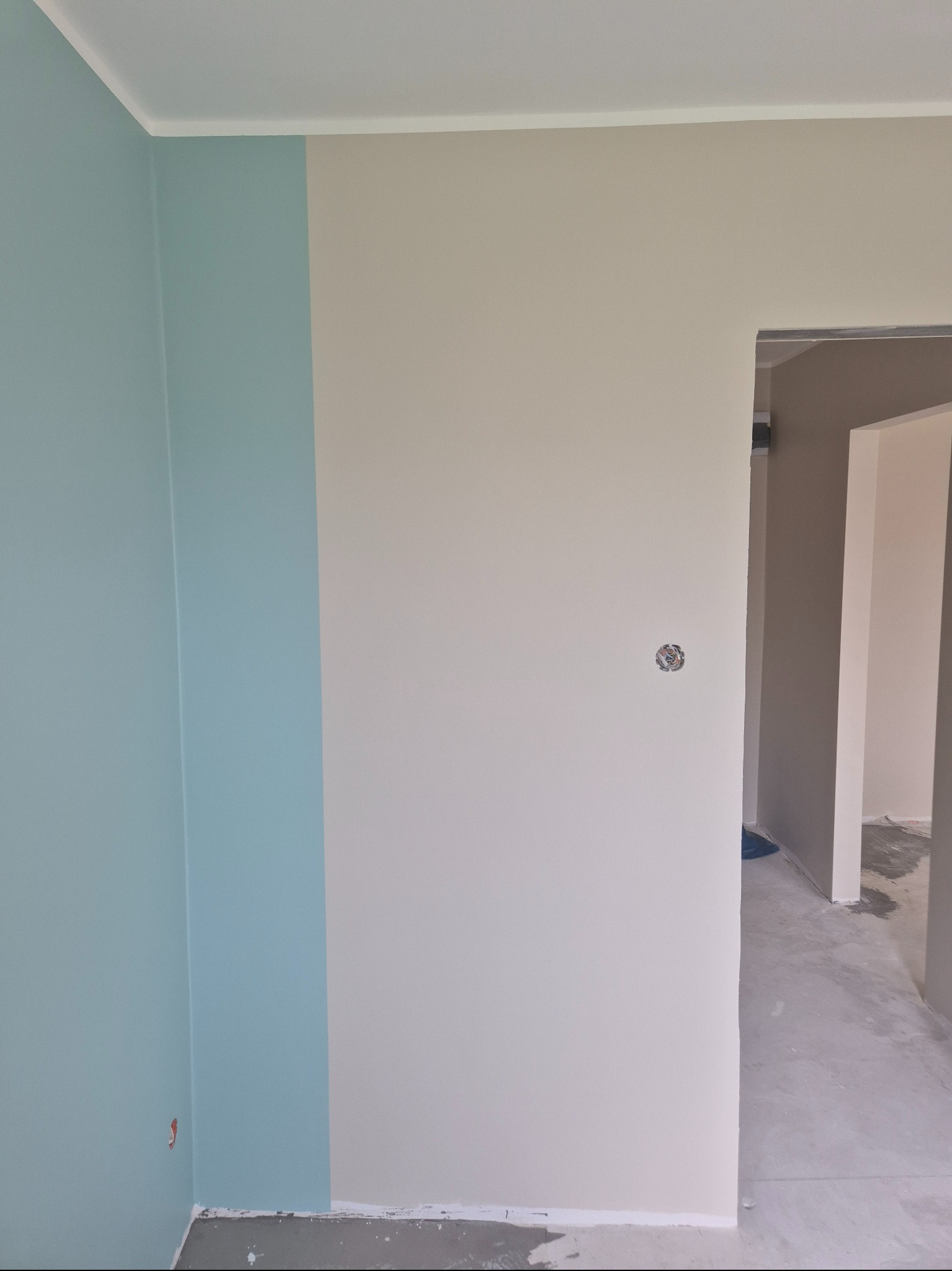 Ściana po malowaniu w odcieniach błękitu i beżu, widoczne gniazdko elektryczne i surowa podłoga w przejściu. Minimalistyczne wykończenie wnętrza.