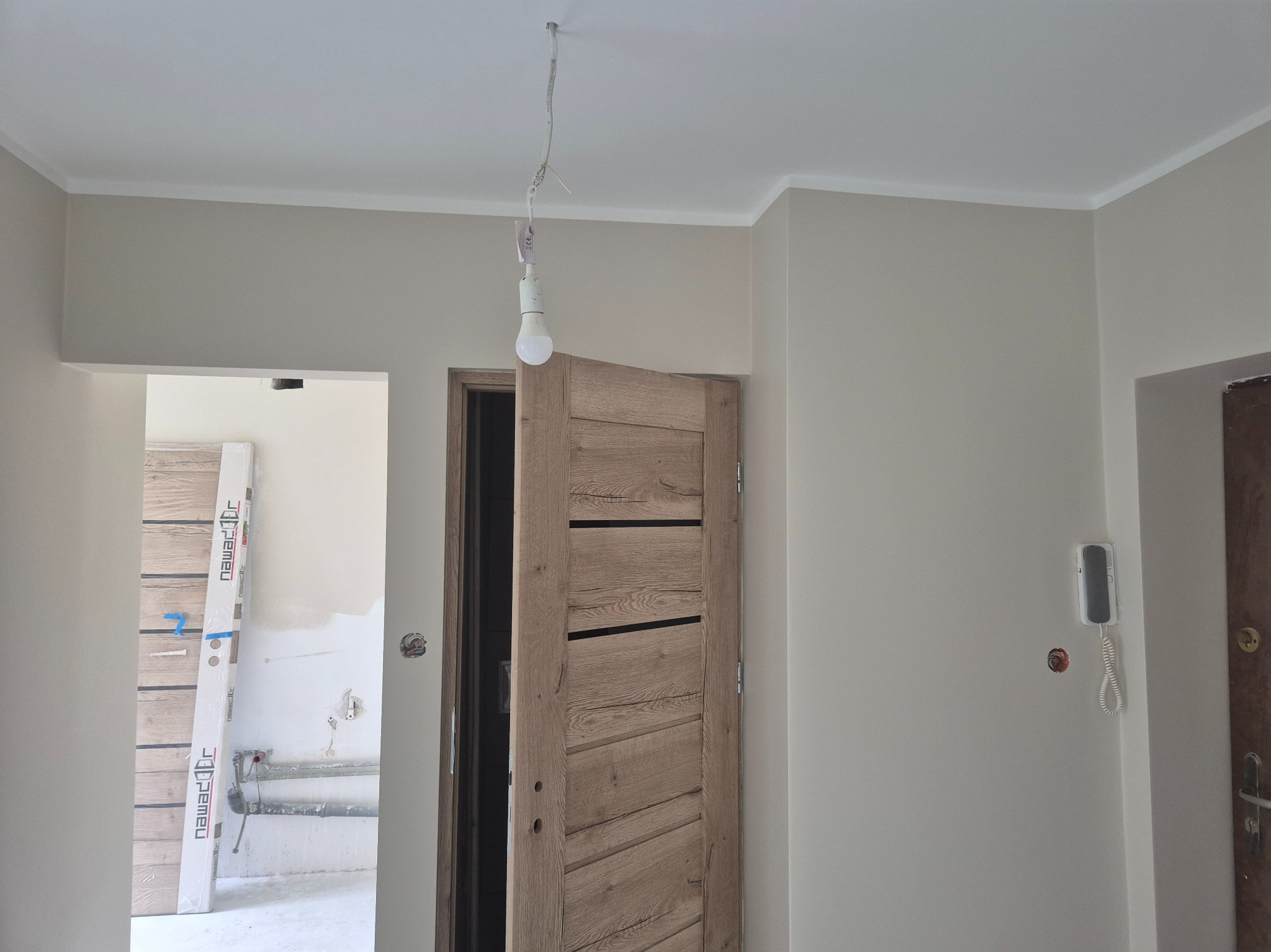 Jasne wnętrze po remoncie: świeżo pomalowane ściany w odcieniach beżu, biały sufit z listwami, nowe drewniane drzwi i widoczne elementy instalacji elektrycznej.