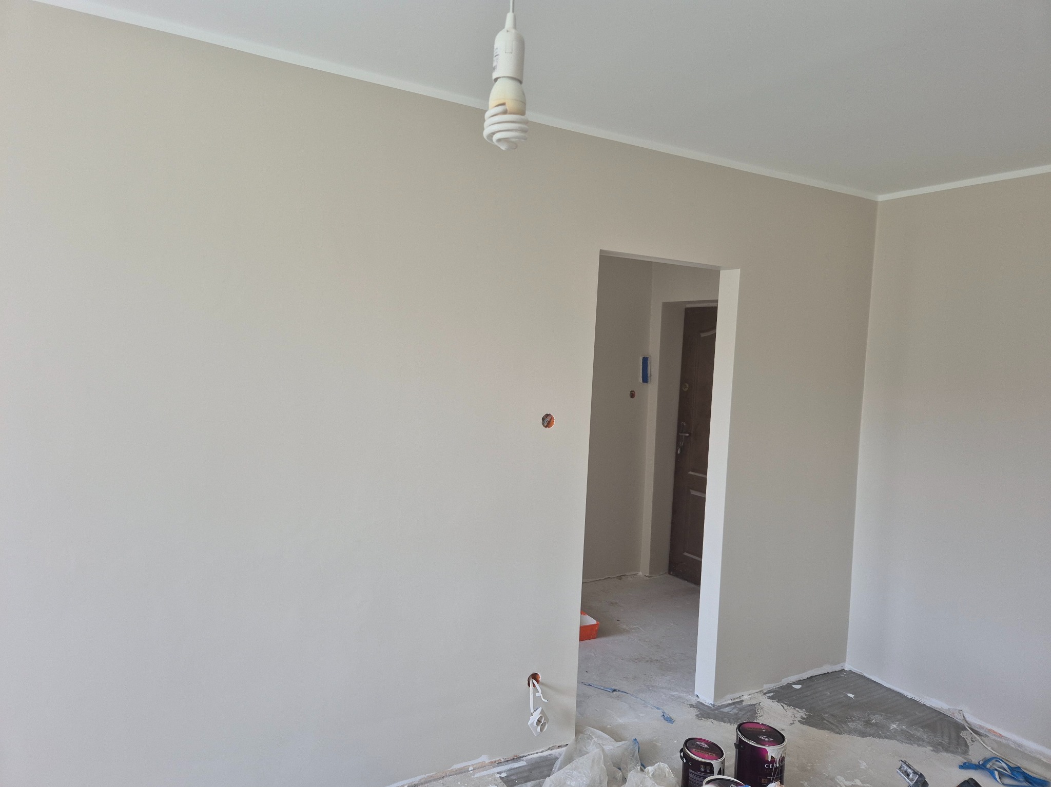 Pomieszczenie po malowaniu ścian na jasny odcień beżu; widoczne puszki farby na podłodze, gniazdka elektryczne bez osłon i listwa przypodłogowa.