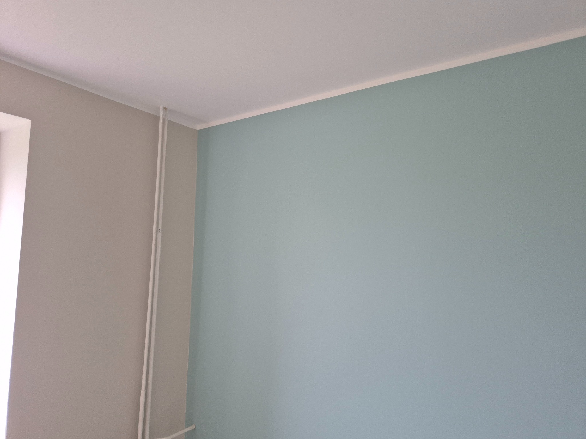 Pomieszczenie po malowaniu: ściany w odcieniach szarości i błękitu, biały sufit z listwą. Dwie rury pionowo przy ścianie. Widok z rogu pokoju.