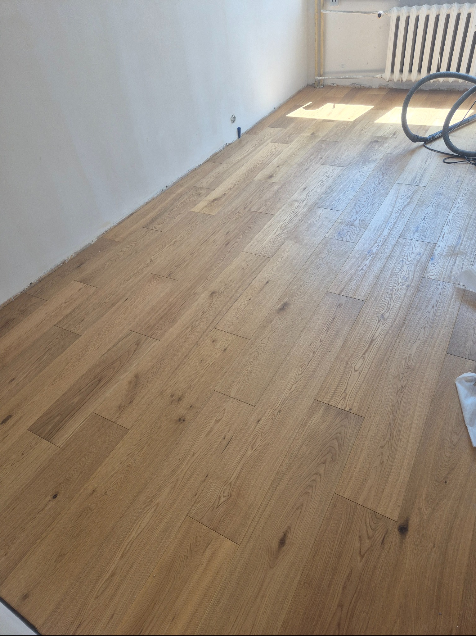 Jasne panele podłogowe ułożone w pokoju z białymi ścianami. Widoczny fragment grzejnika i rura od odkurzacza. Podłoga po ułożeniu, gotowa do użytku.