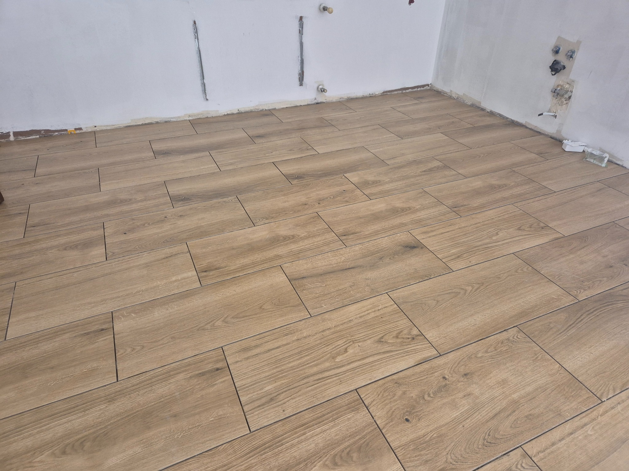 Ukończona podłoga z płytek imitujących drewno w surowym wnętrzu. Widoczne niedokończone ściany i wystające instalacje. Jasne, naturalne światło.