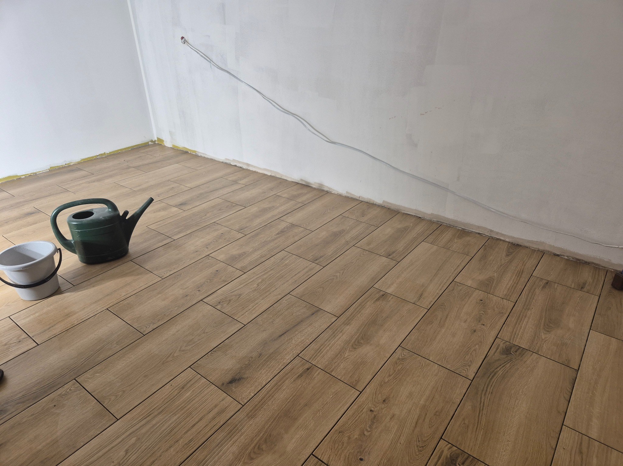 Ułożone panele podłogowe w odcieniach brązu, białe ściany w trakcie malowania, widoczne kable i akcesoria malarskie: konewka i wiadro.
