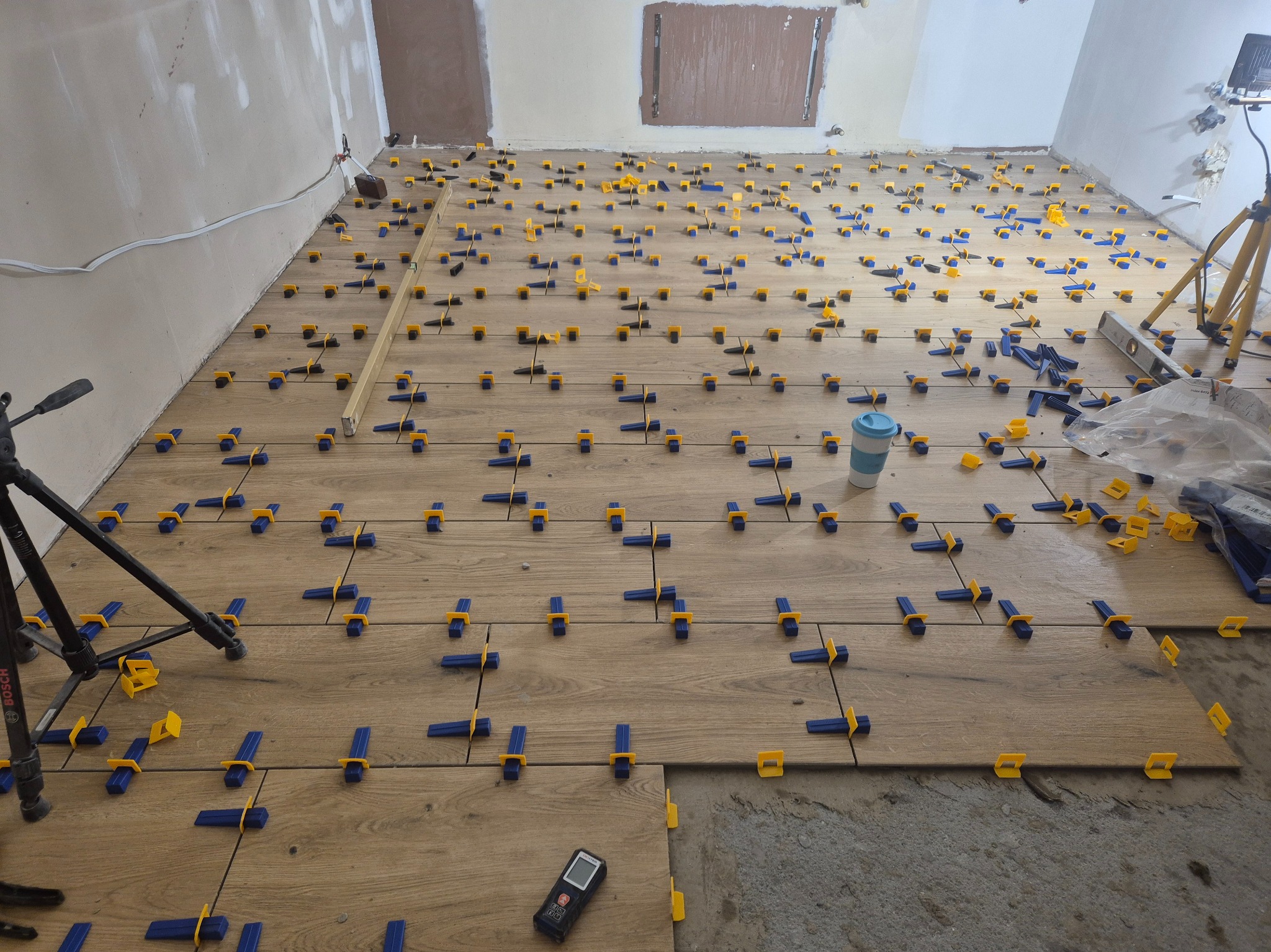 Układanie płytek podłogowych z użyciem systemu poziomowania: niebieskie kliny i żółte klipsy na drewnopodobnych płytkach. Widoczny laserowy miernik odległości.