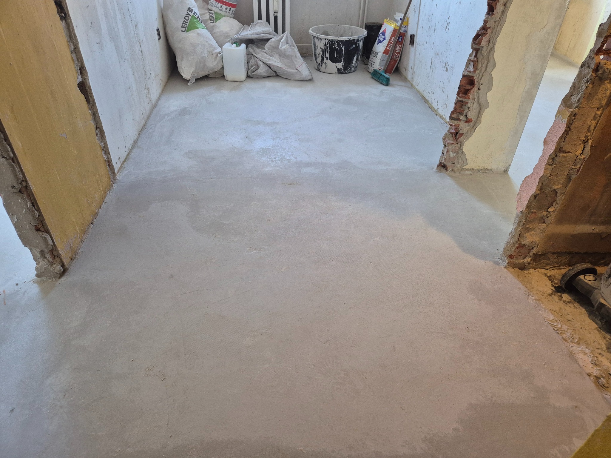 Wyrównana, szara posadzka w remontowanym pomieszczeniu z widocznymi workami z zaprawą i narzędziami w tle. Fragmenty ścian odsłonięte z cegieł.
