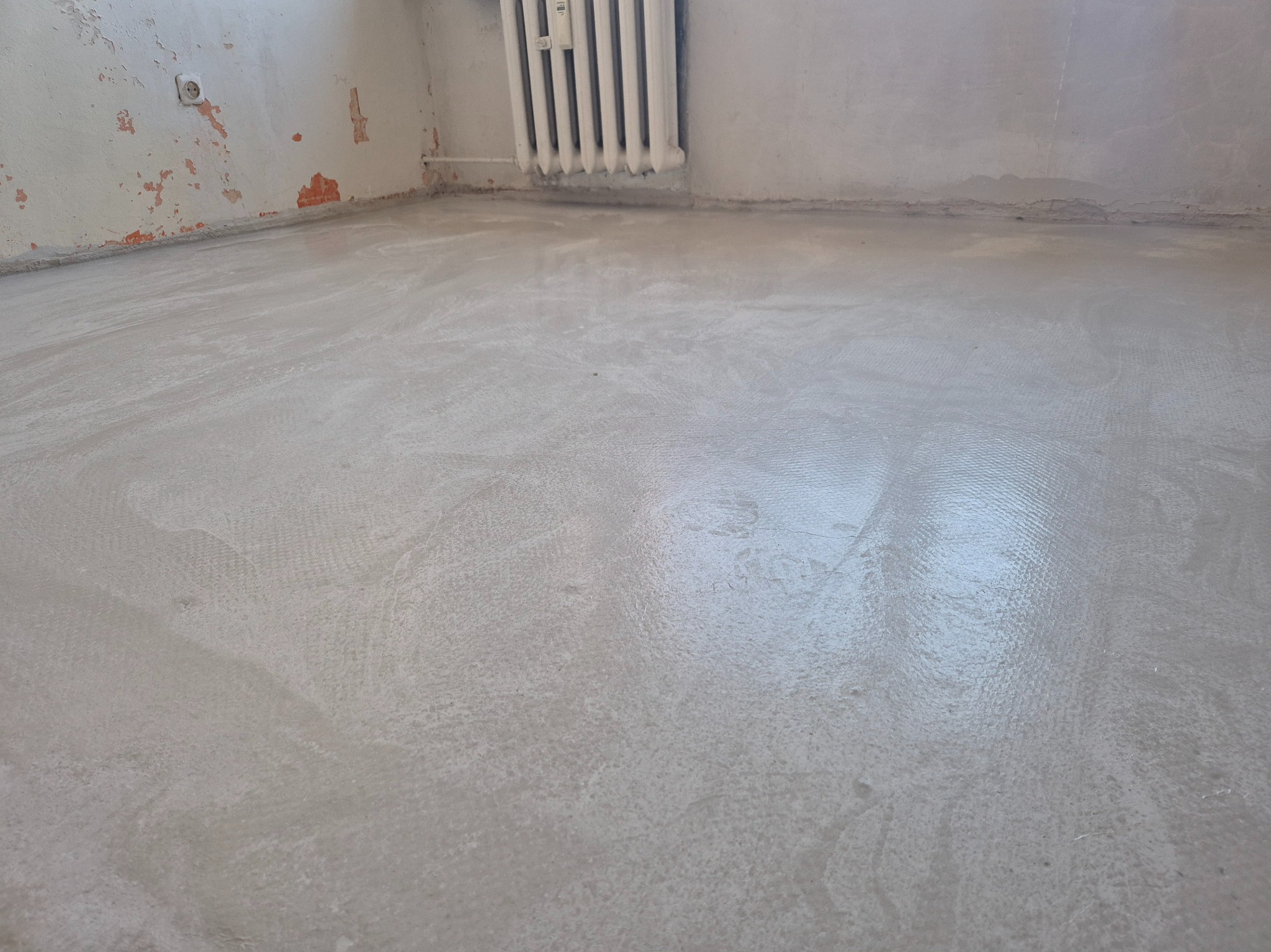 Wyrównana, szara wylewka podłogowa w pokoju z grzejnikiem i fragmentami starej farby na ścianach. Widoczne ślady po wałku na powierzchni.
