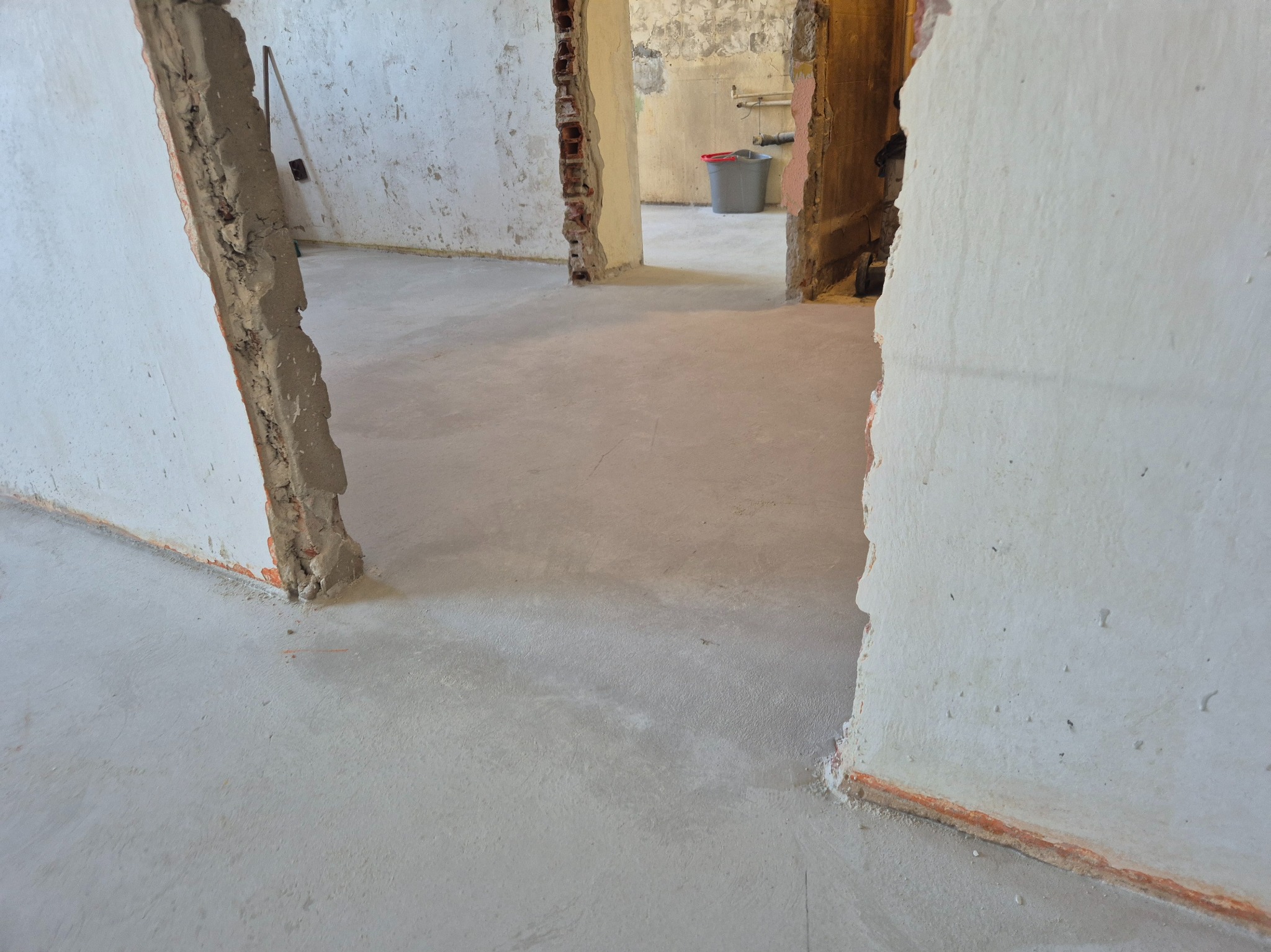 Wnętrze w trakcie prac wykończeniowych: surowa podłoga, ściany z fragmentami cegieł i resztkami tynku, widoczne przejścia do innych pomieszczeń.