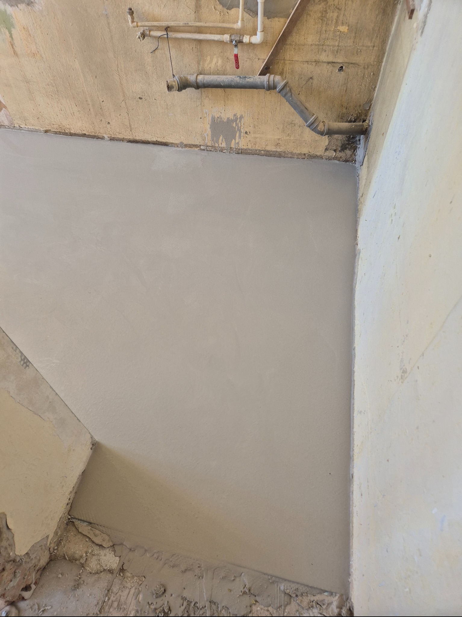 Wyrównana, świeżo wylana szara posadzka w rogu pomieszczenia z widocznymi rurami pod sufitem. Ściany w trakcie remontu.