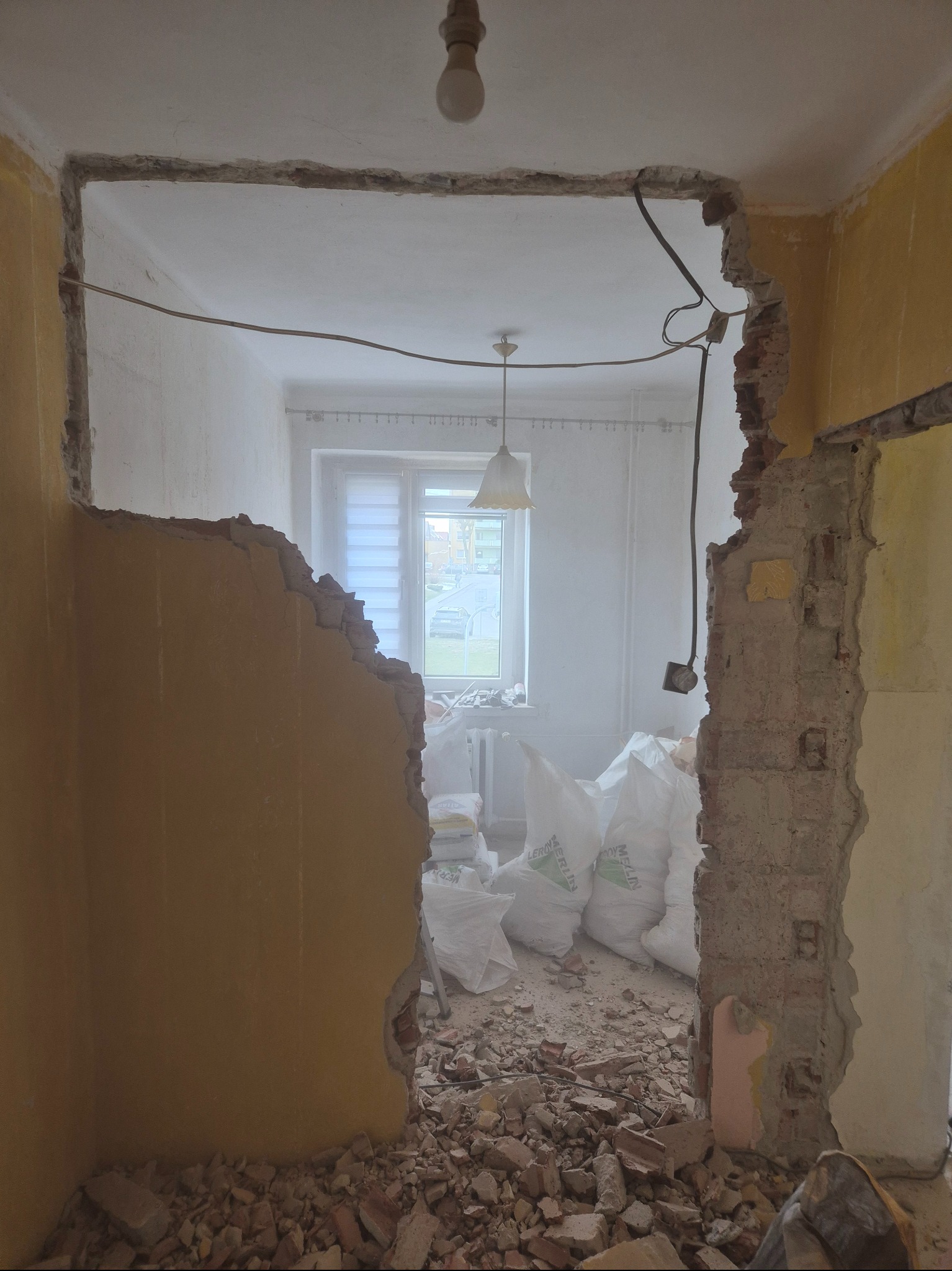Demontaż ściany działowej z cegły w mieszkaniu. Widoczne worki z gruzem, odsłonięte przewody elektryczne i fragmenty starej instalacji. Żółte ściany i resztki tapet.