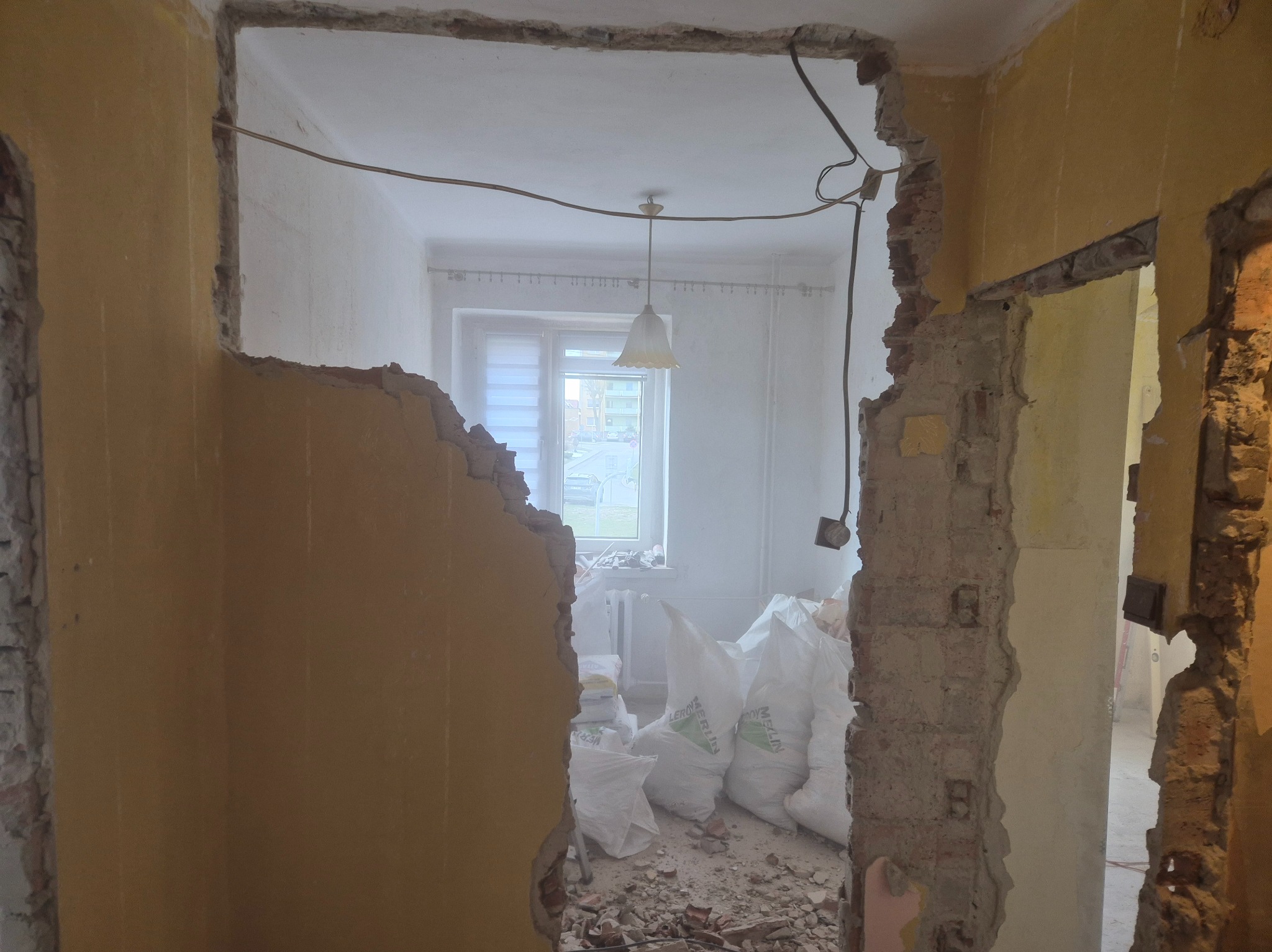 Wnętrze w trakcie prac remontowych: zniszczone ściany, widoczne warstwy muru, worki z materiałami budowlanymi w tle, okno z widokiem na zewnątrz.
