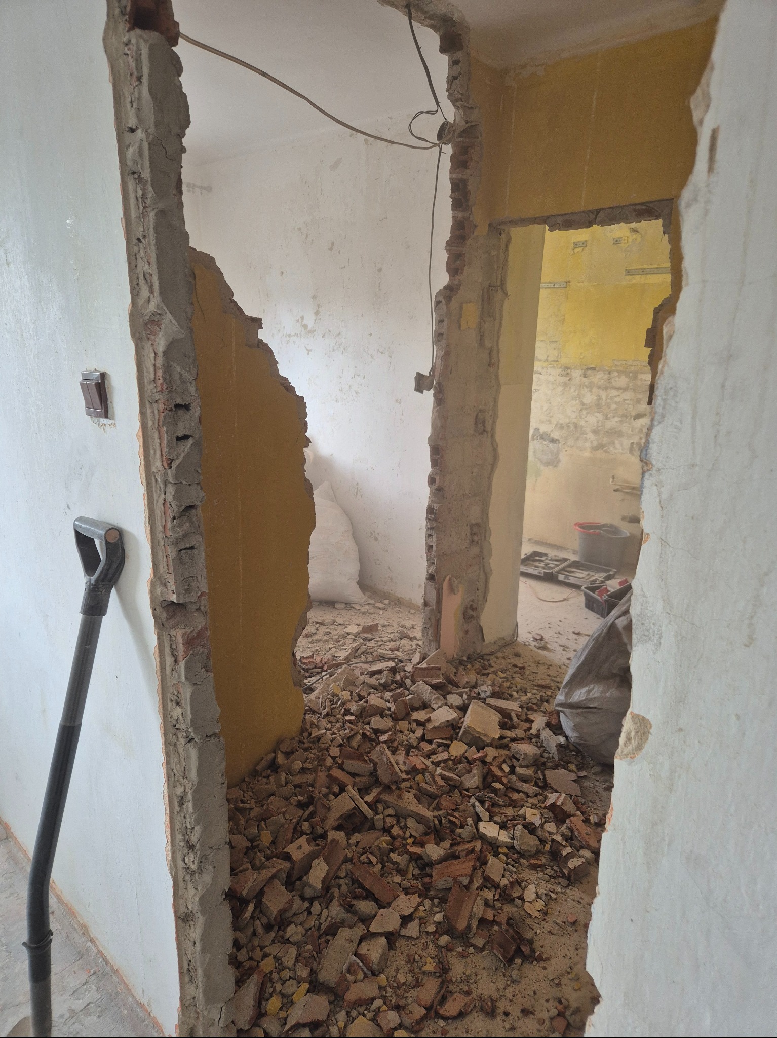 Demontaż ścian z cegły podczas remontu. Gruz ceglany na podłodze, widoczne fragmenty instalacji elektrycznej. Łopata oparta o ścianę.