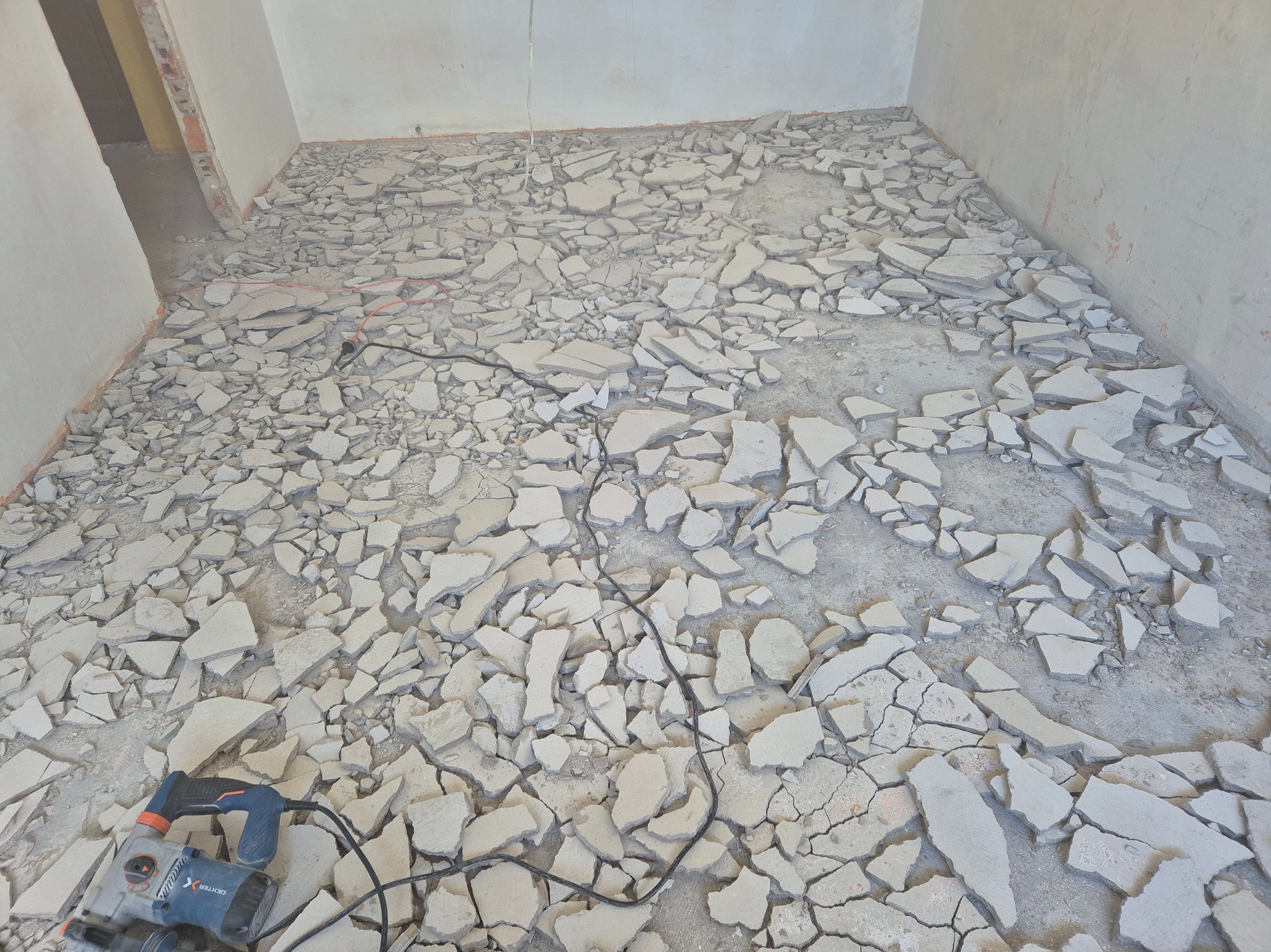 Demontaż starej podłogi w pokoju: gruz i fragmenty posadzki pokrywają podłoże. Widoczny młot udarowy i kabel zasilający. Ściany w surowym stanie.