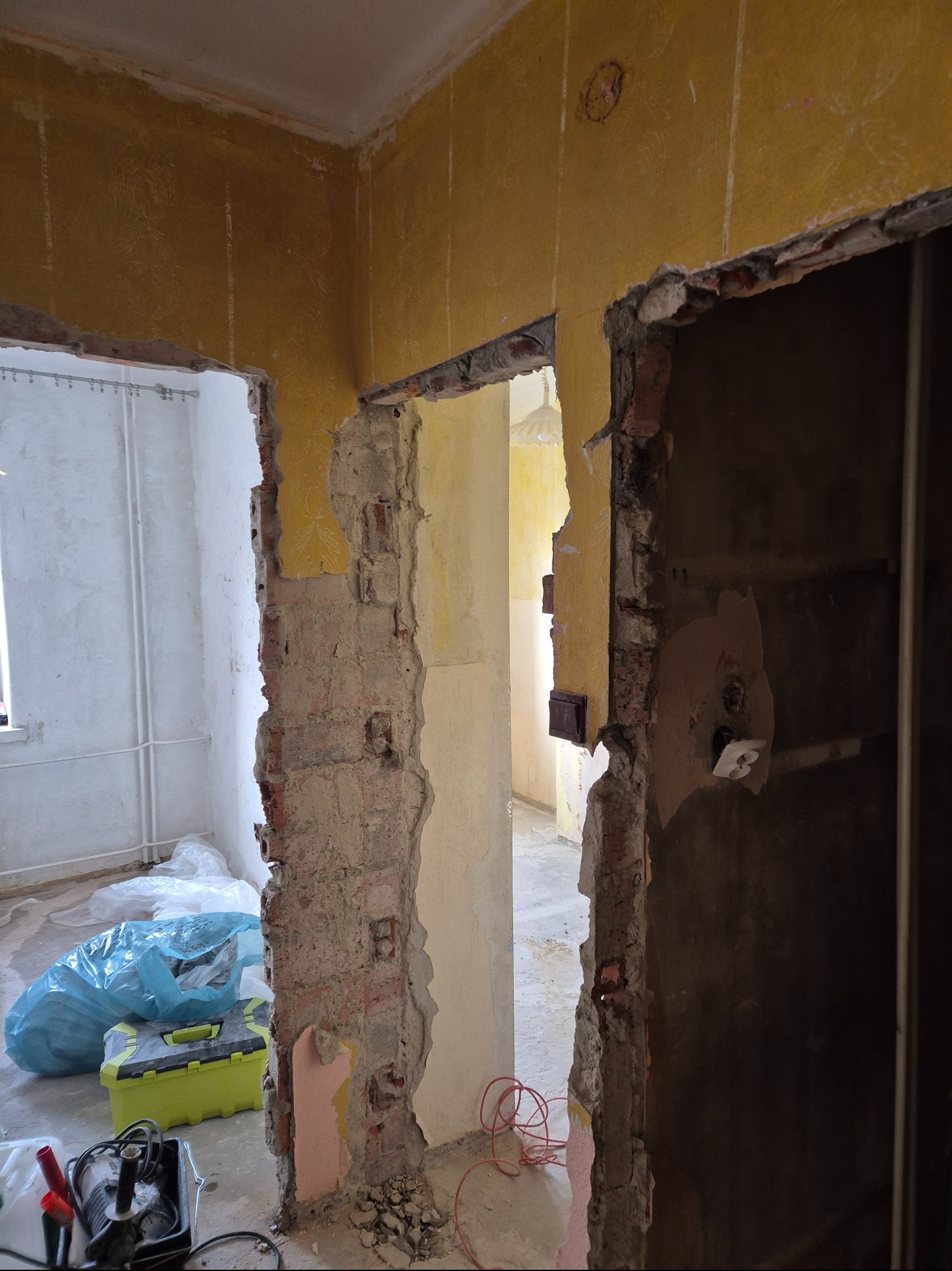 Wnętrze w trakcie demontażu: odsłonięte cegły, resztki tapet na żółtej ścianie, widoczne kable i narzędzia, sugerujące przygotowania do remontu.
