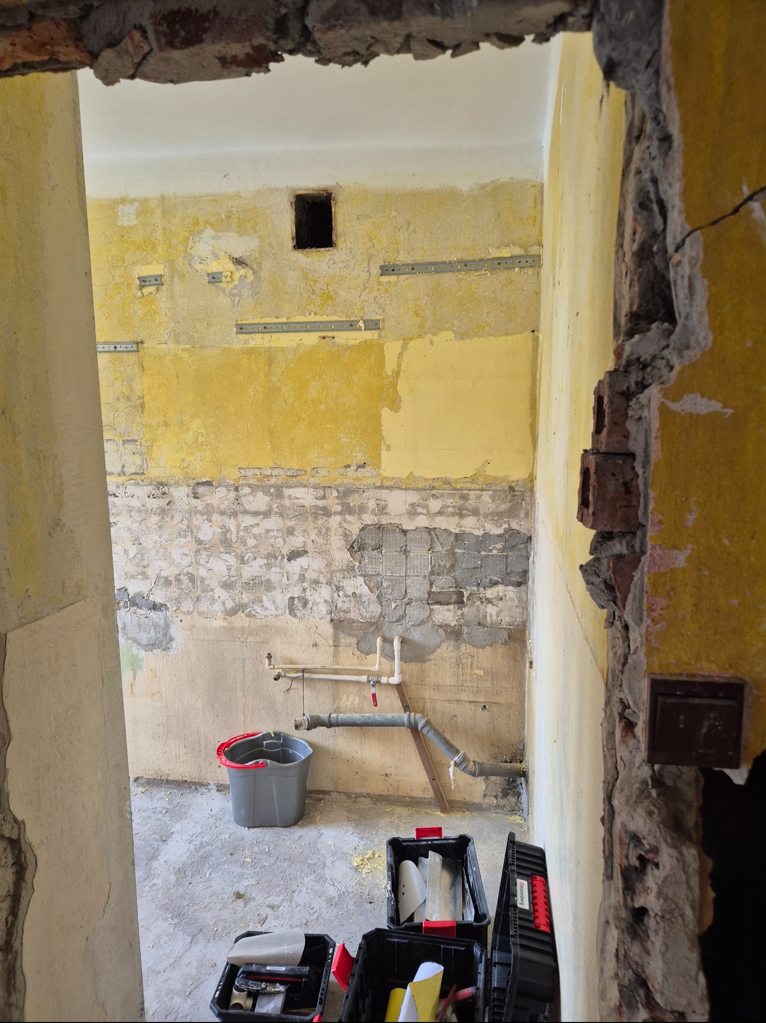 Wnętrze w trakcie remontu: odsłonięte ściany z fragmentami starej farby, instalacja wodna, narzędzia w skrzynkach i gruz. Widok przez zdemontowane futryny drzwi.