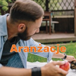 Gardea - Mężczyzna w dżinsowej koszuli układa owoce na kocu piknikowym przed drewnianą altaną w ogrodzie.