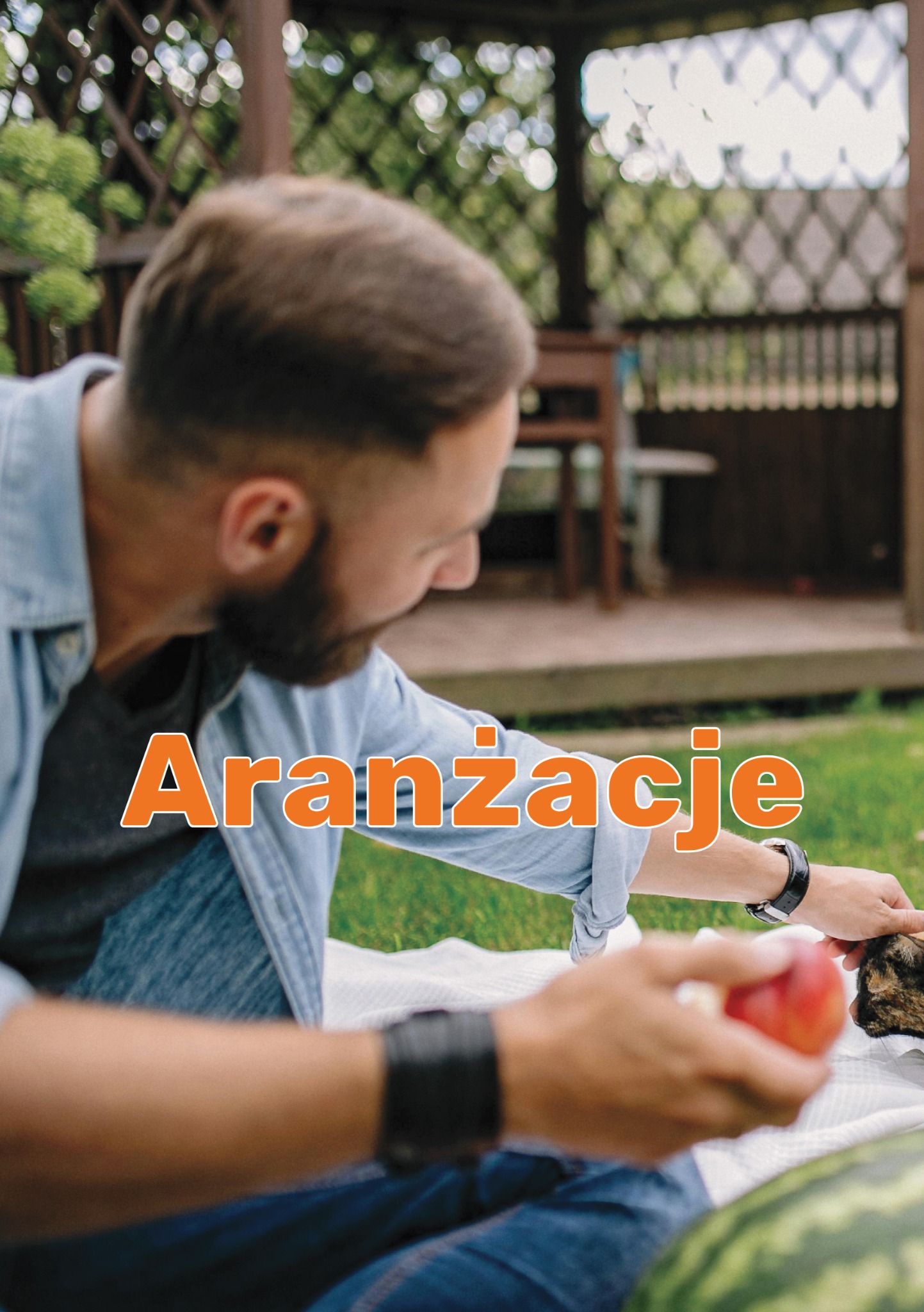 Mężczyzna w dżinsowej koszuli układa owoce na kocu piknikowym przed drewnianą altaną w ogrodzie.