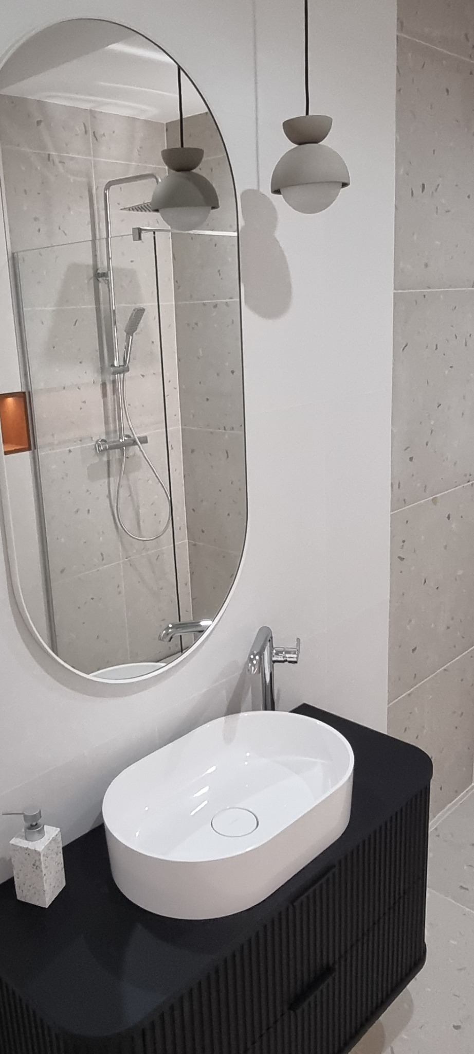 Nowoczesna łazienka: owalne lustro odbija prysznic, biała umywalka na czarnej szafce z żłobieniami, minimalistyczne lampy wiszące i płytki imitujące lastryko.