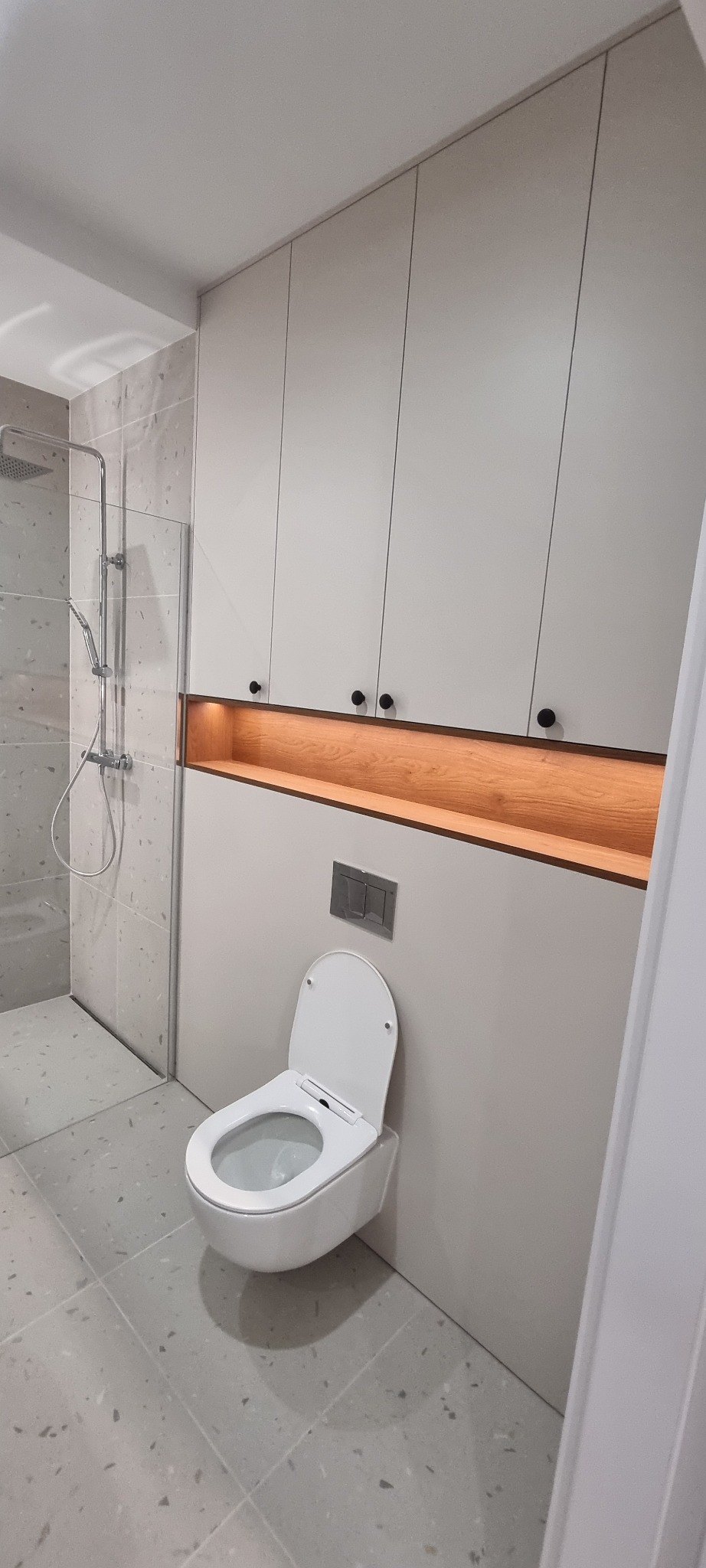 Nowoczesna łazienka z podwieszaną toaletą, szafkami w zabudowie i prysznicem typu walk-in. Szare płytki podłogowe i ścienne, oświetlenie LED w niszy.